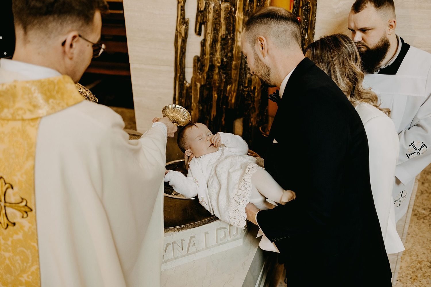 Catholic priest pours holy water over infant during baptism sacrament at church font.