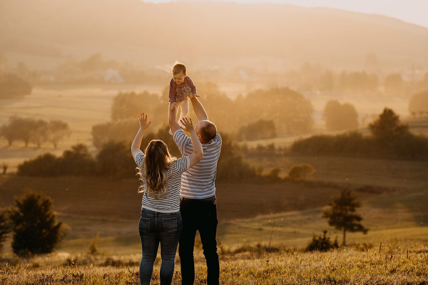 Parents lifting baby in golden sunset light over scenic countryside meadow.