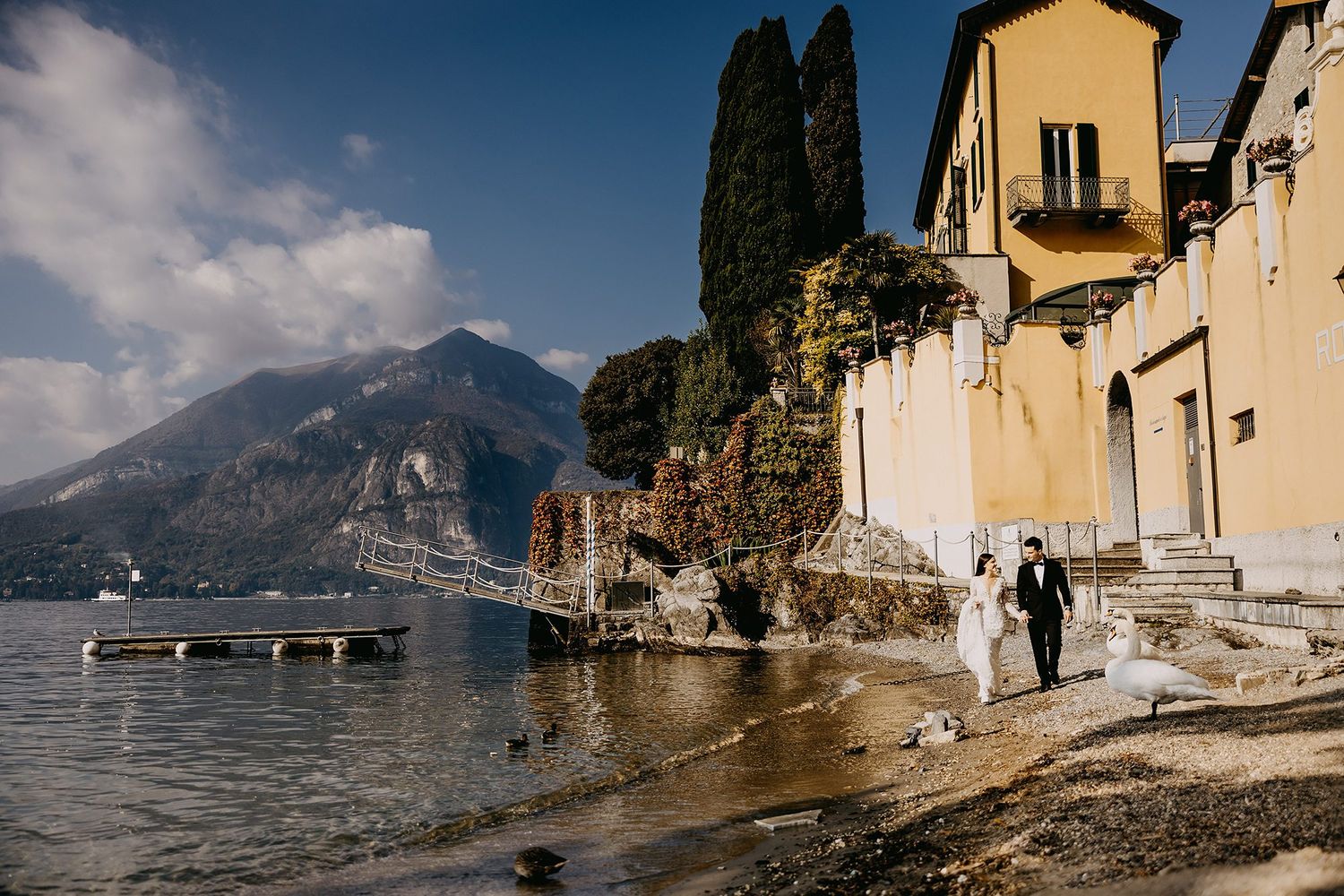 Wedding couple walking along Lake Como shoreline with stunning Italian villa and mountain backdrop.