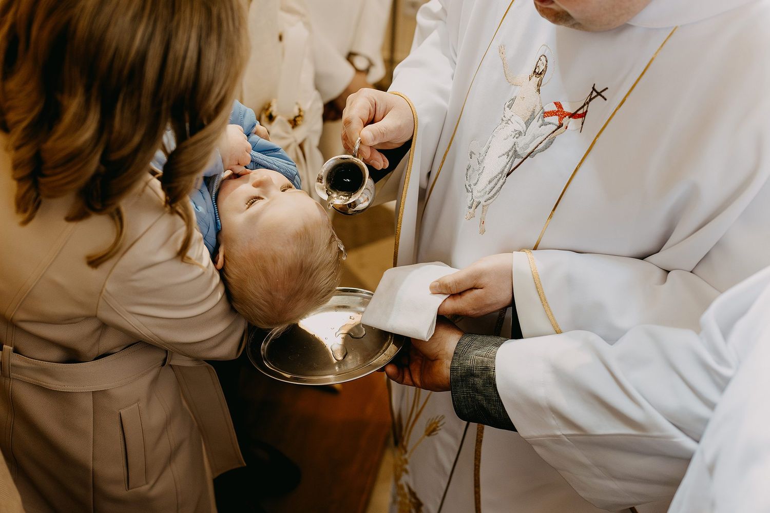 Catholic priest performing infant baptism ceremony, pouring holy water over babys head at baptismal font.