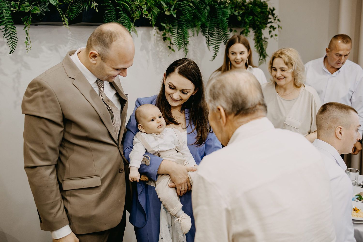 A woman in blue holds a baby while an elderly man and family gather at a christening.