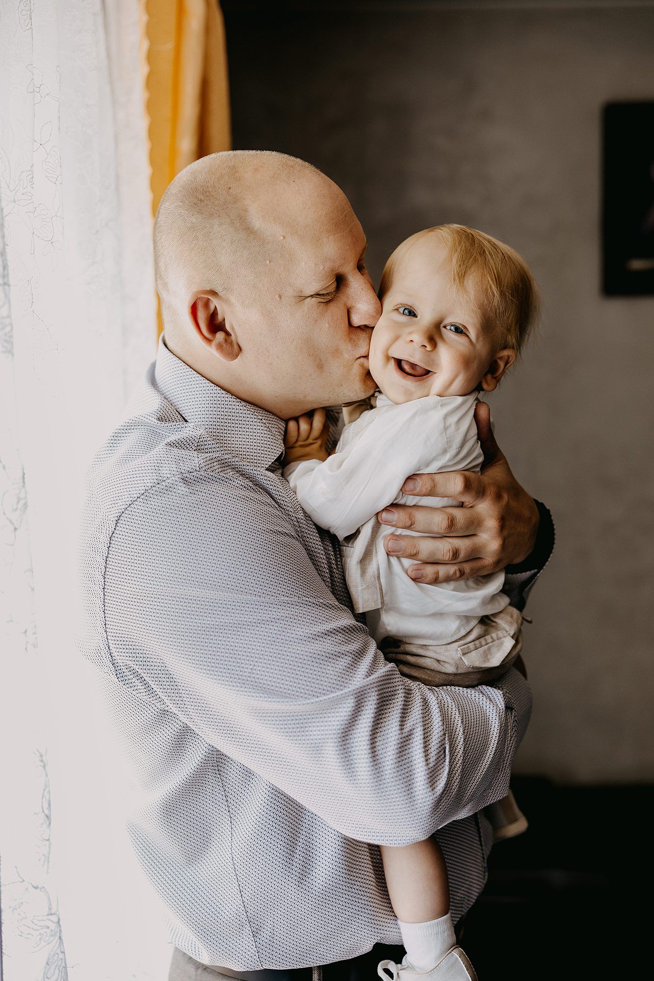 Bald father tenderly kissing his laughing baby while holding the child in his arms indoors.