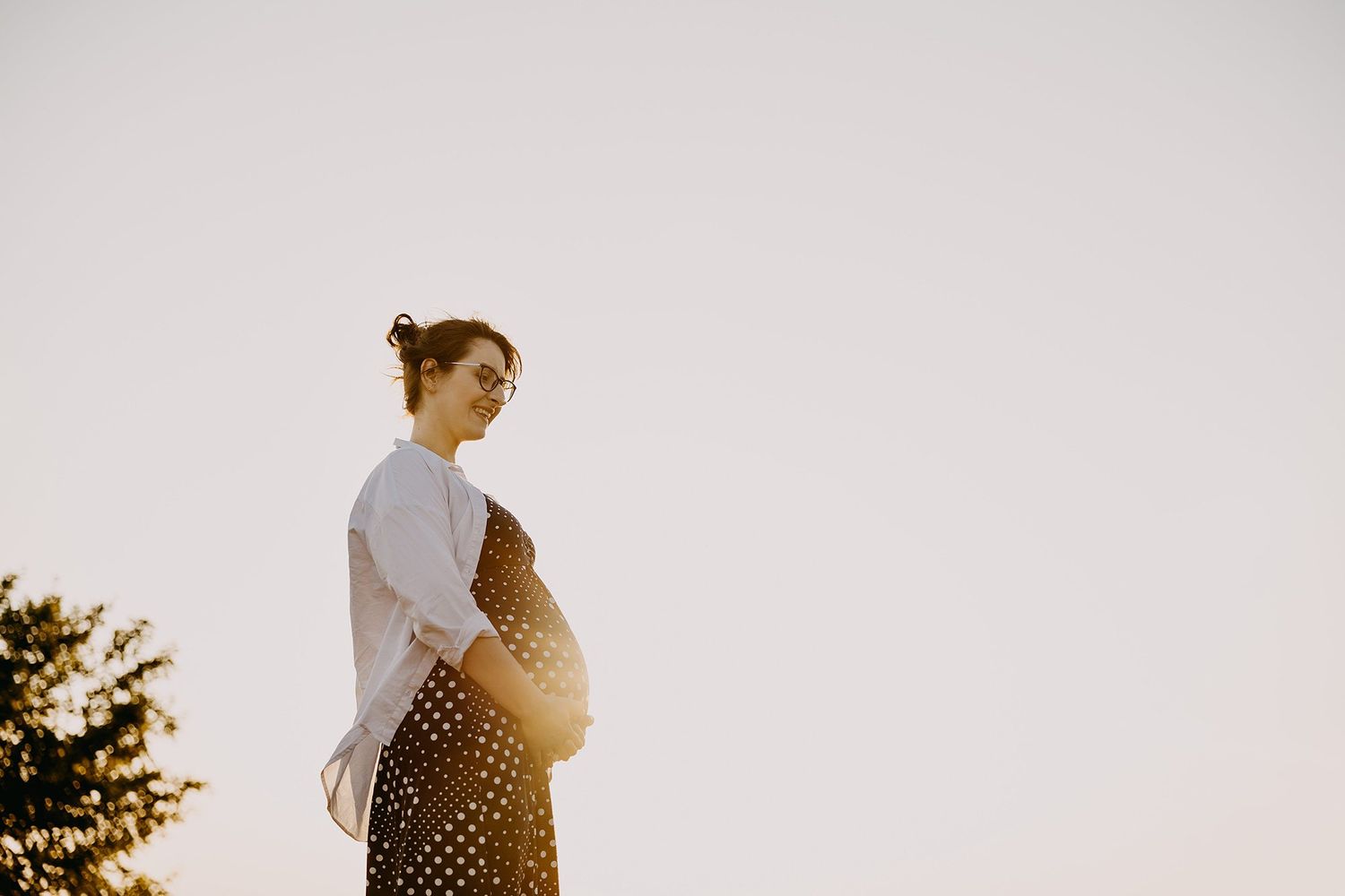 Pregnant woman in white shirt and polka dot skirt cradling bump against bright sky at sunset.