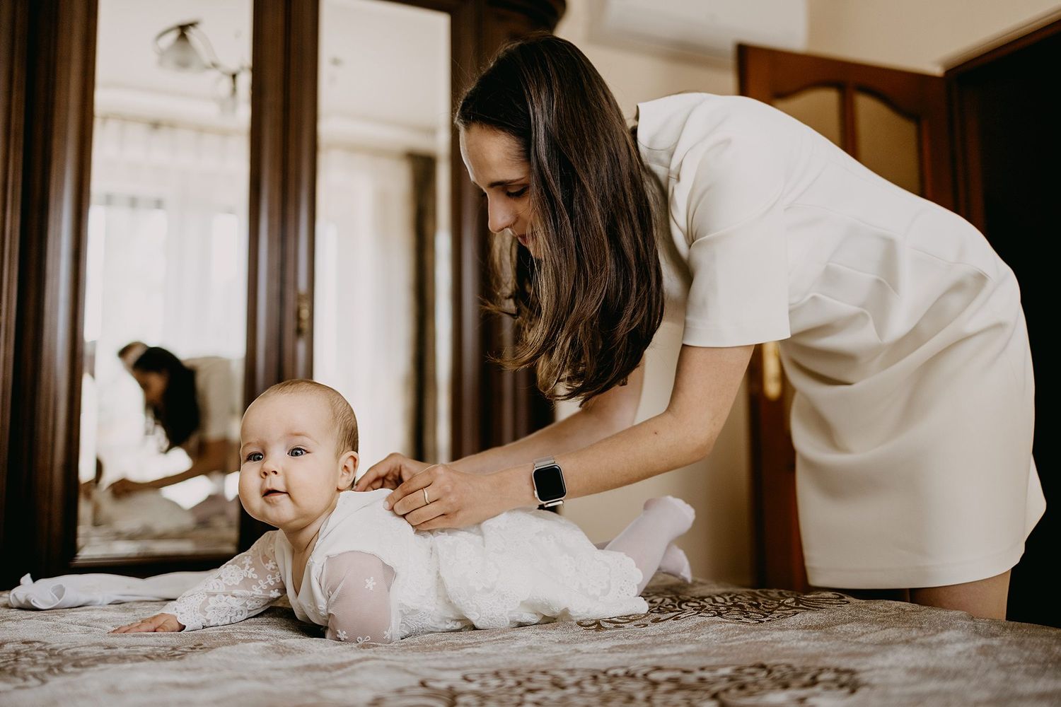 Young mother in white dress dressing her baby on a bed in an elegant room.
