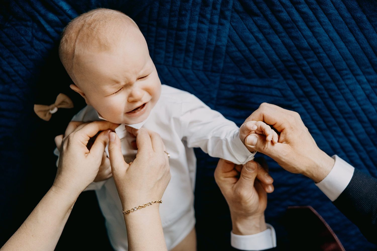 Newborn baby in white outfit being dressed by parents hands on blue blanket.