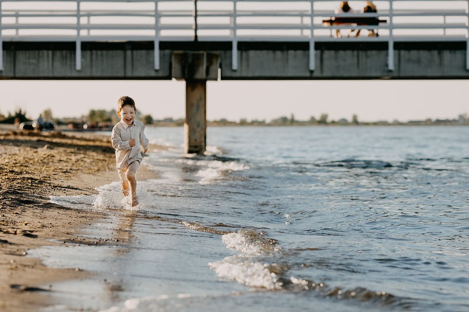 Young child runs joyfully along sandy shoreline with waves splashing near a pier.
