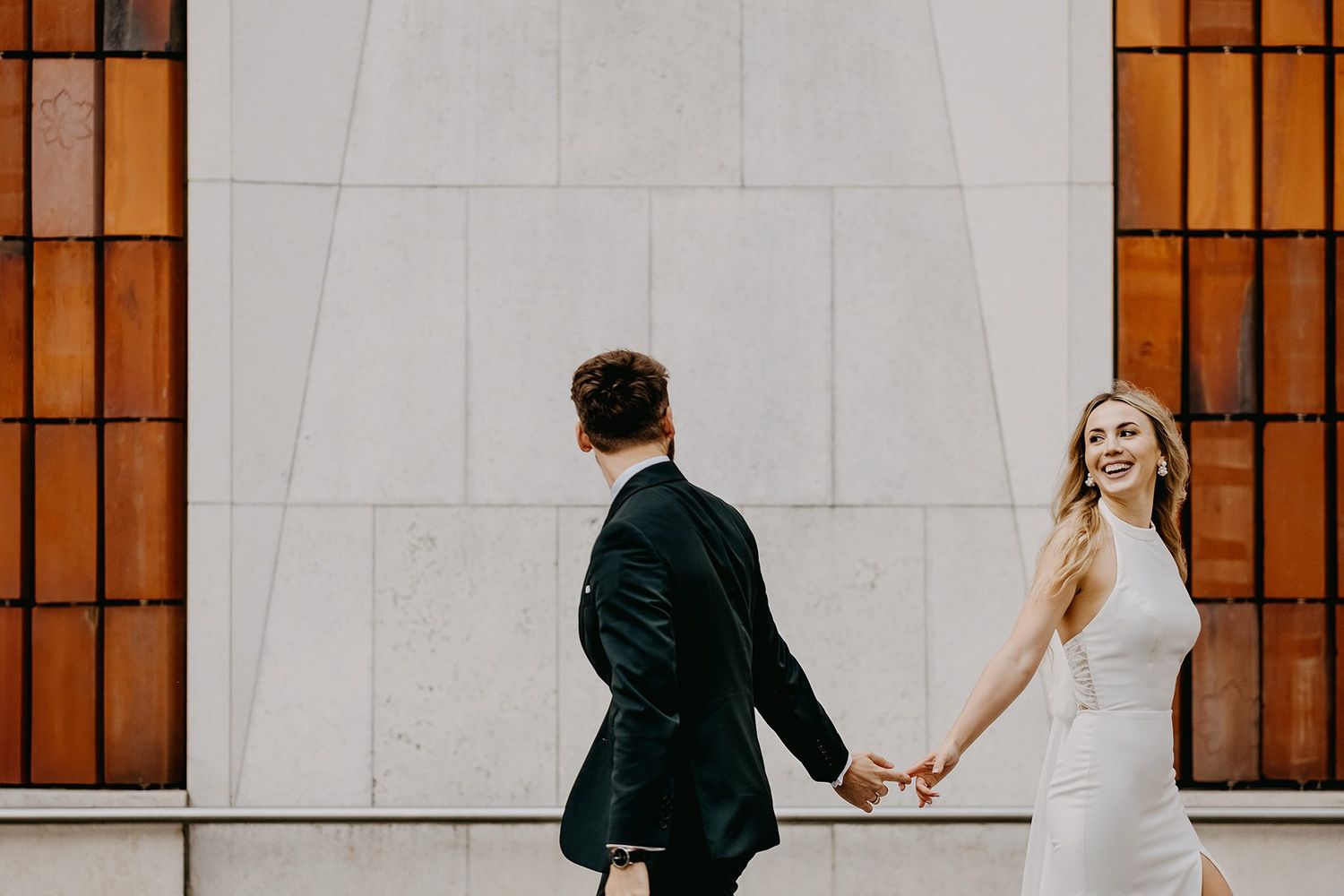 A smiling bride in a white dress holds hands with a groom in a black suit walking past a tiled wall.