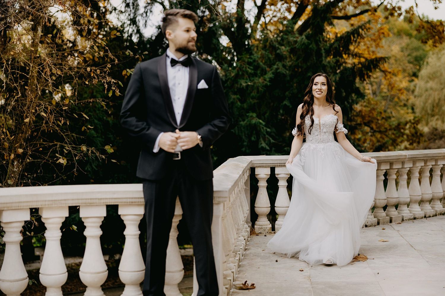 Bride in flowing white gown and groom in black tuxedo pose on elegant stone balustrade surrounded by autumn foliage.