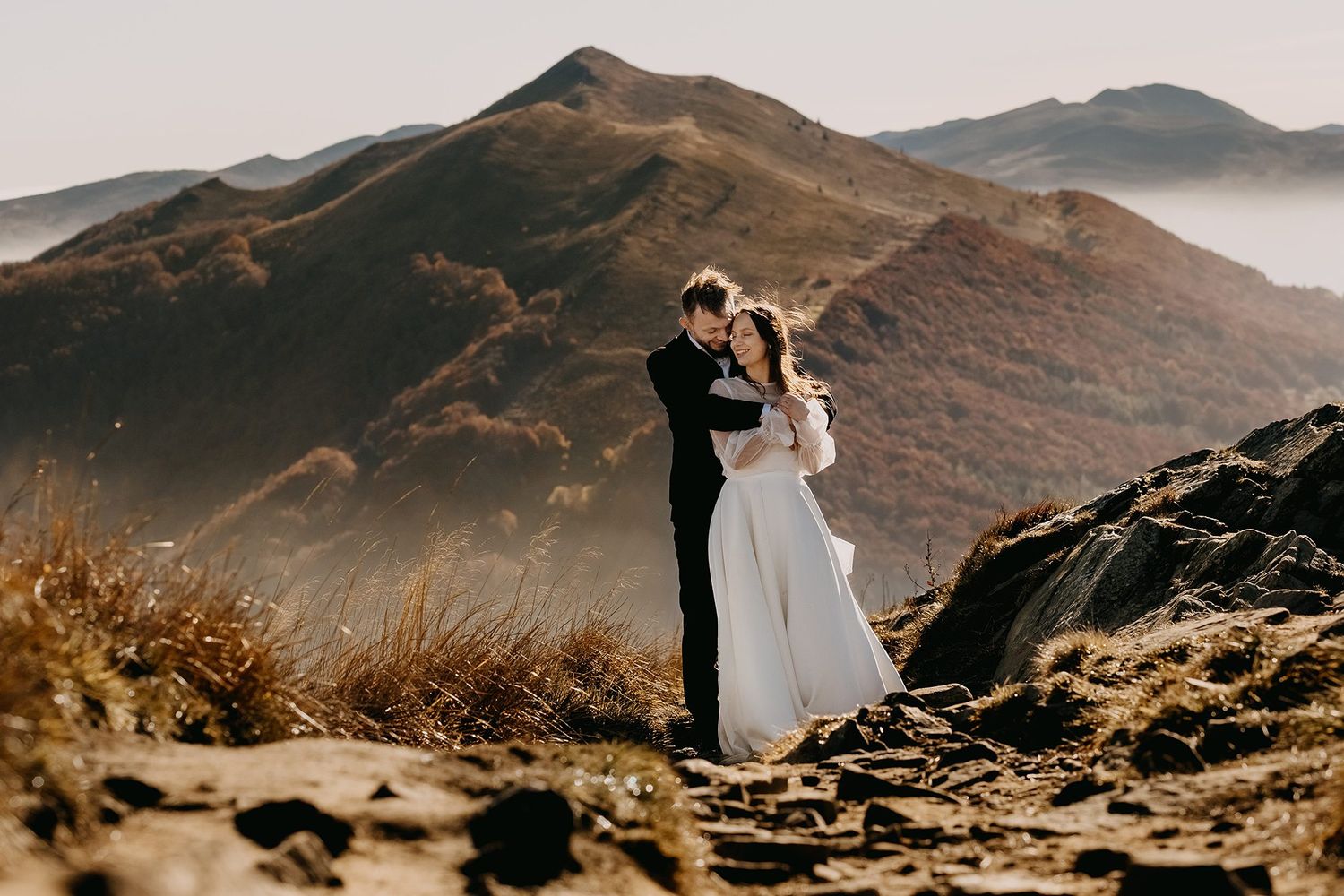 Bride and groom embracing on a rocky mountain summit with golden autumn hills in the background.