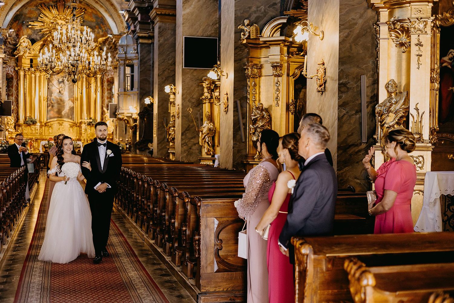 Bride and groom walking down the aisle of an ornate golden baroque church with guests watching from the pews.
