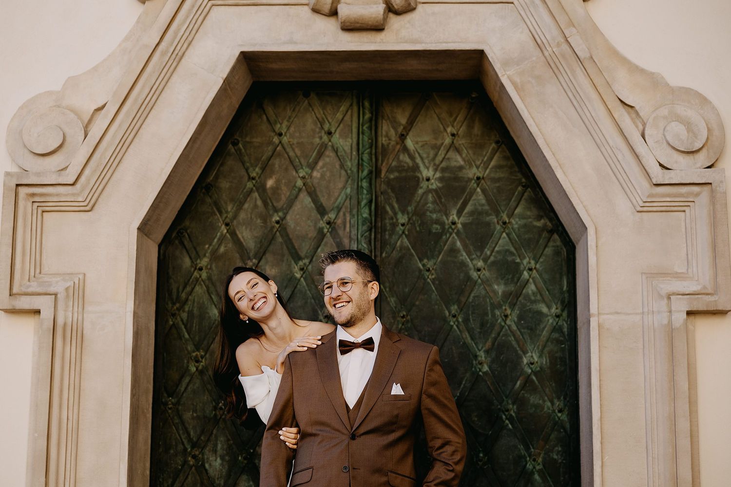 Smiling bride and groom pose before an ornate green antique door in elegant wedding attire.