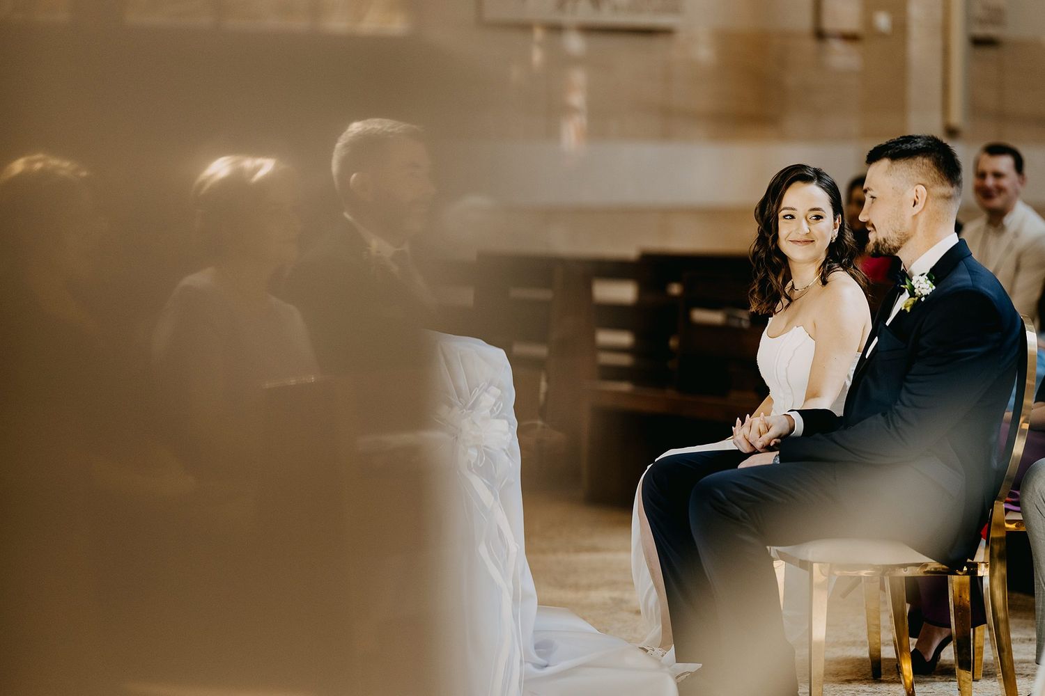 Bride and groom smiling warmly at each other during a sunlit wedding ceremony.