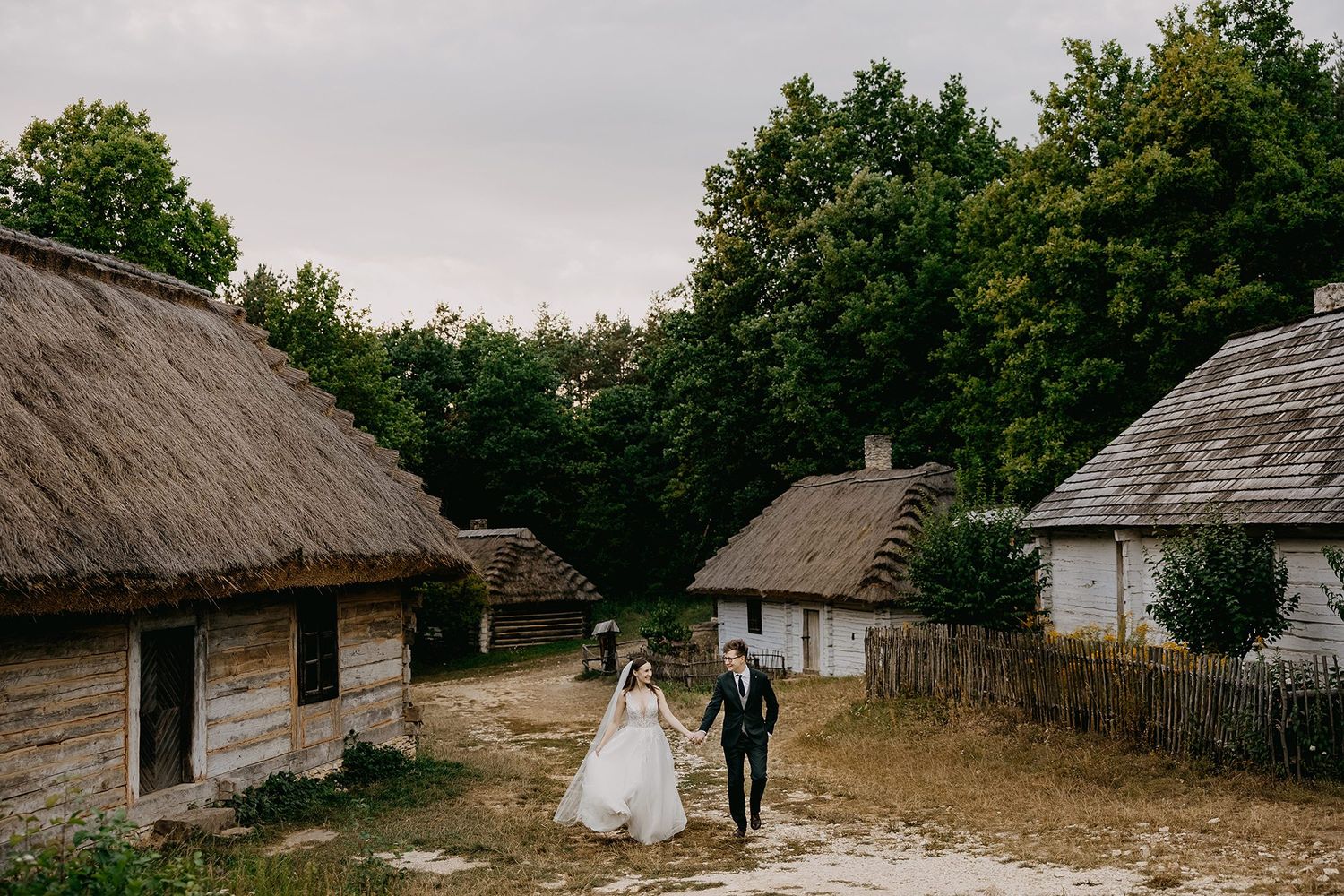Bride and groom walking through a rustic village with thatched-roof cottages and trees.