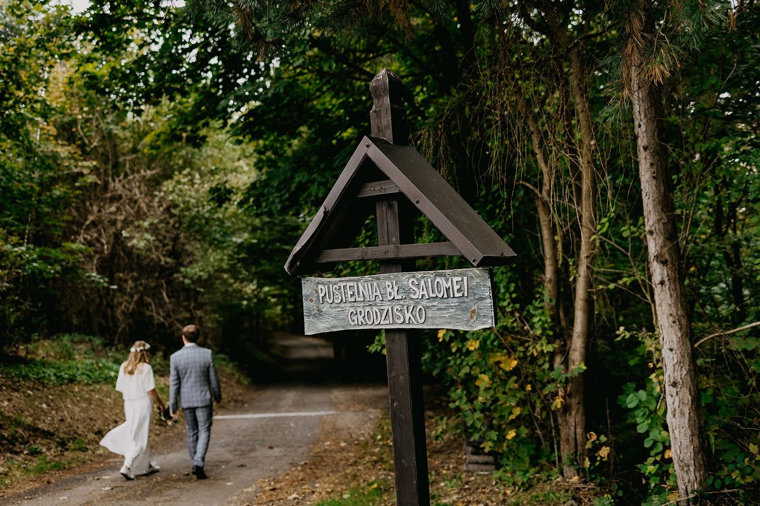 Bride and groom walk down forest path past rustic wooden wedding sign.