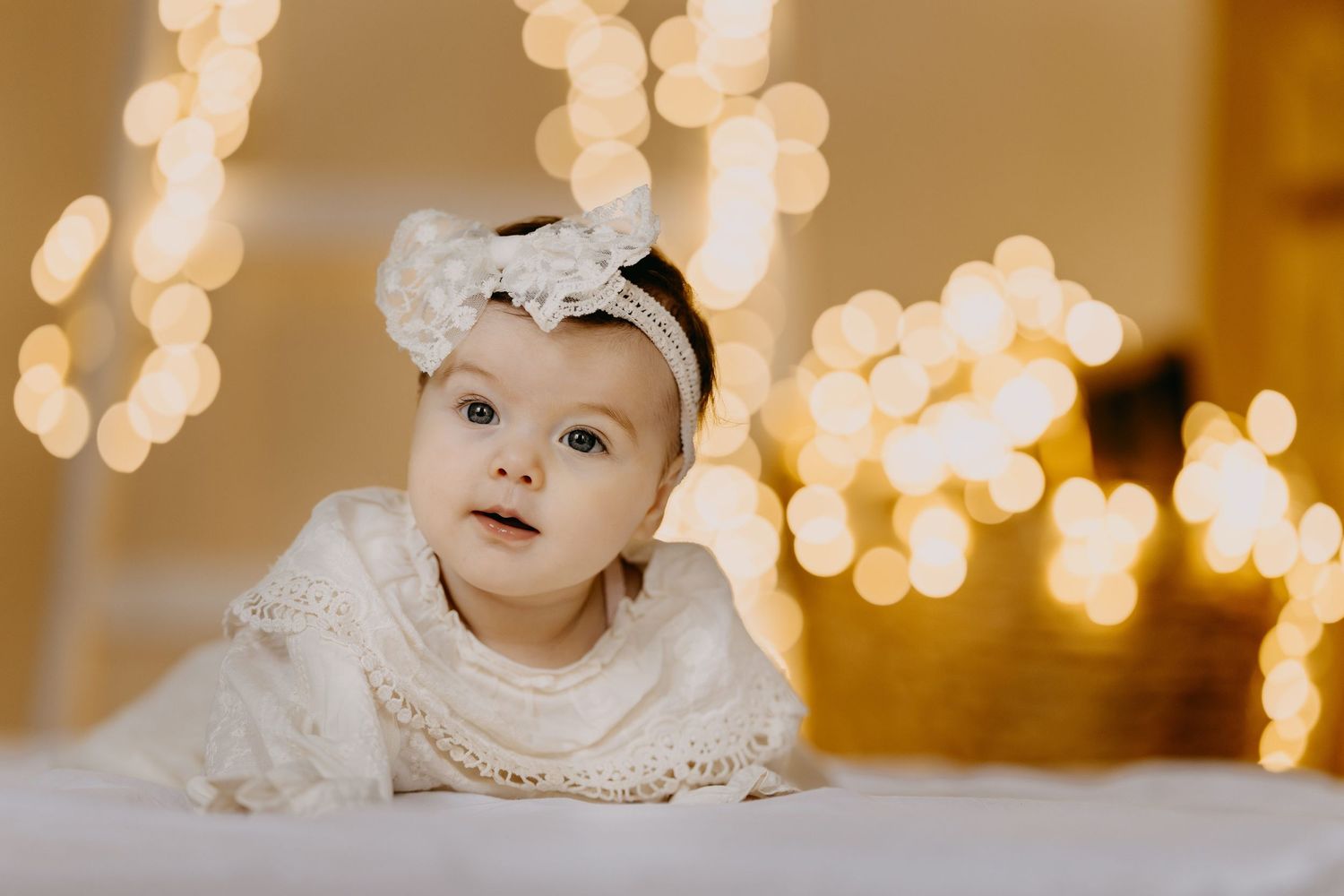Baby girl in a white lace dress and floral headband lying on her tummy against a warm background of glowing golden bokeh lights.