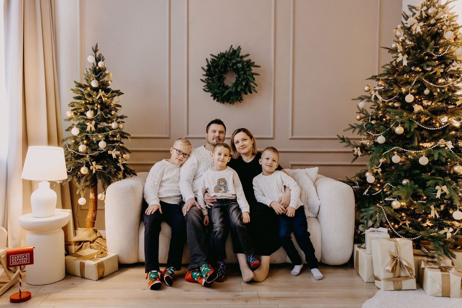 Family of five in matching white tops poses on a cozy sofa between two decorated Christmas trees with gift boxes and a wreath on the wall.