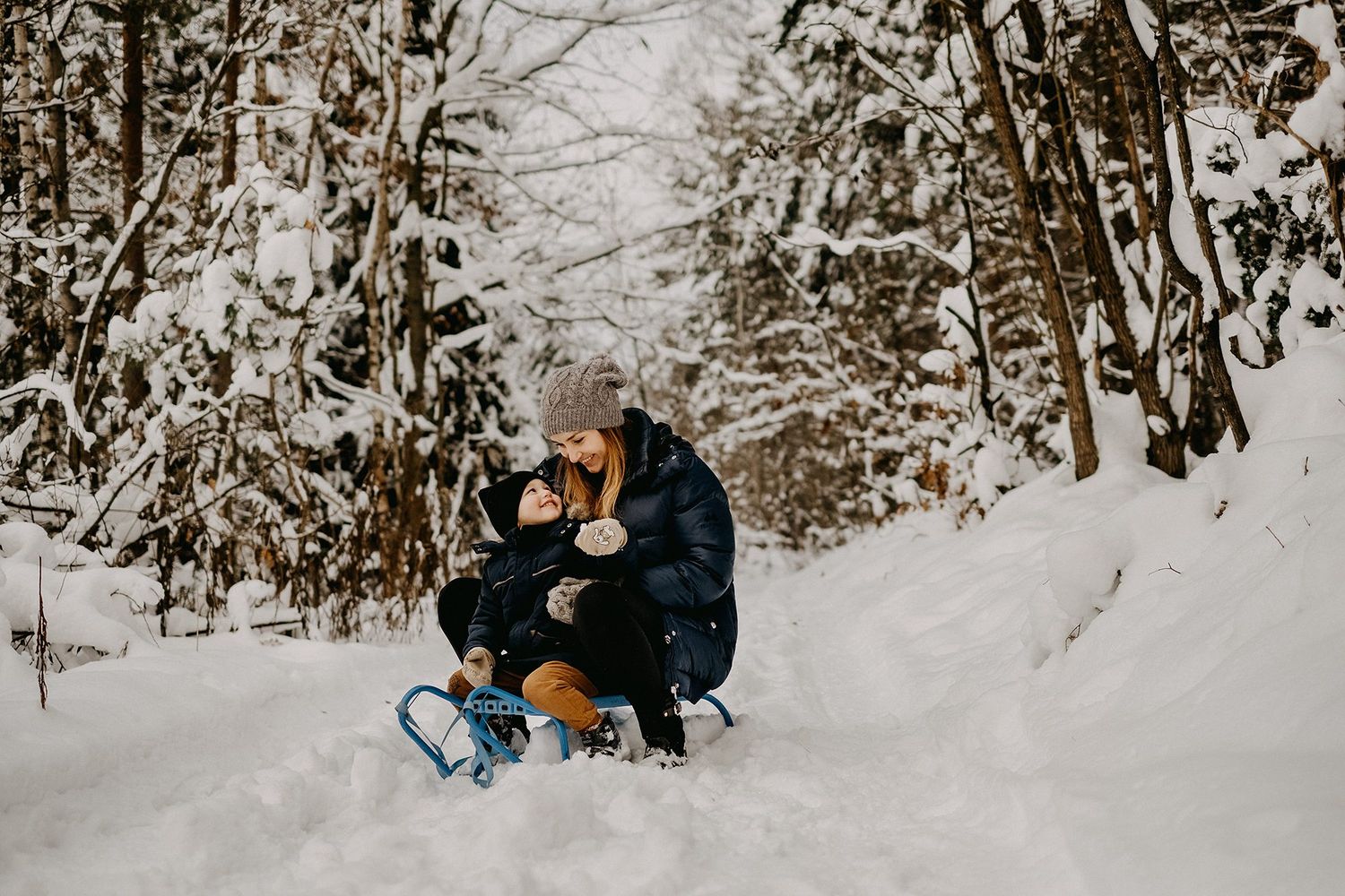 Mother and child sledding together on snowy forest trail in winter wonderland setting.