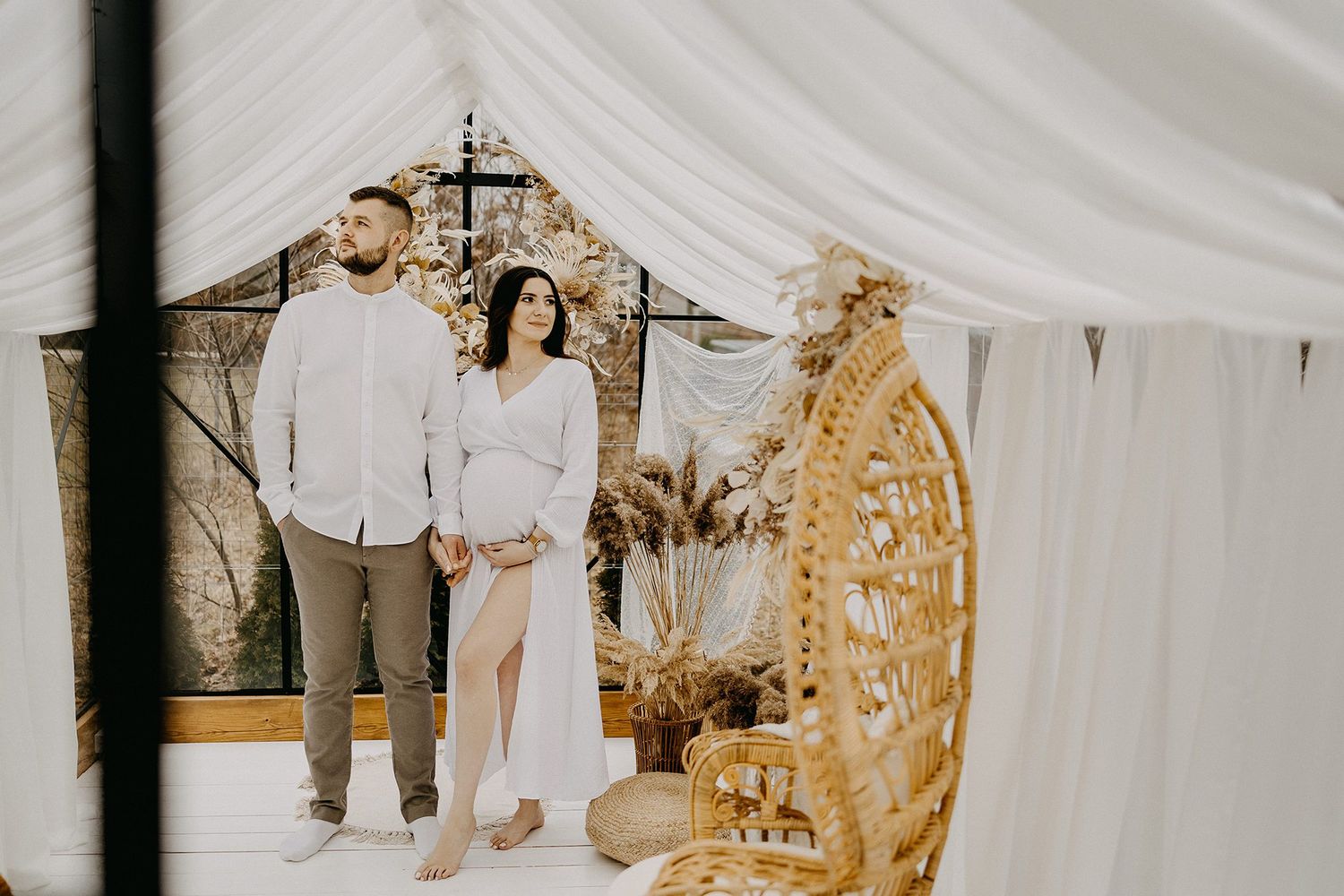 Expectant couple holding hands inside elegant white draped tent with wicker chair decor.