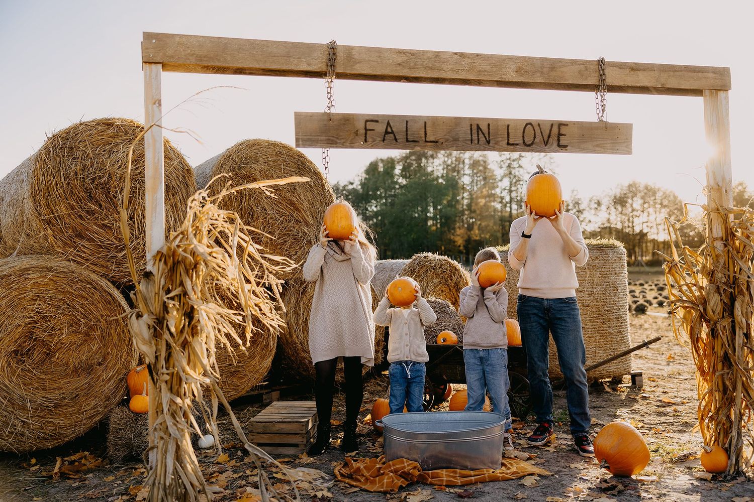 Family at a fall pumpkin patch holding pumpkins in front of a Fall In Love sign with hay bales.