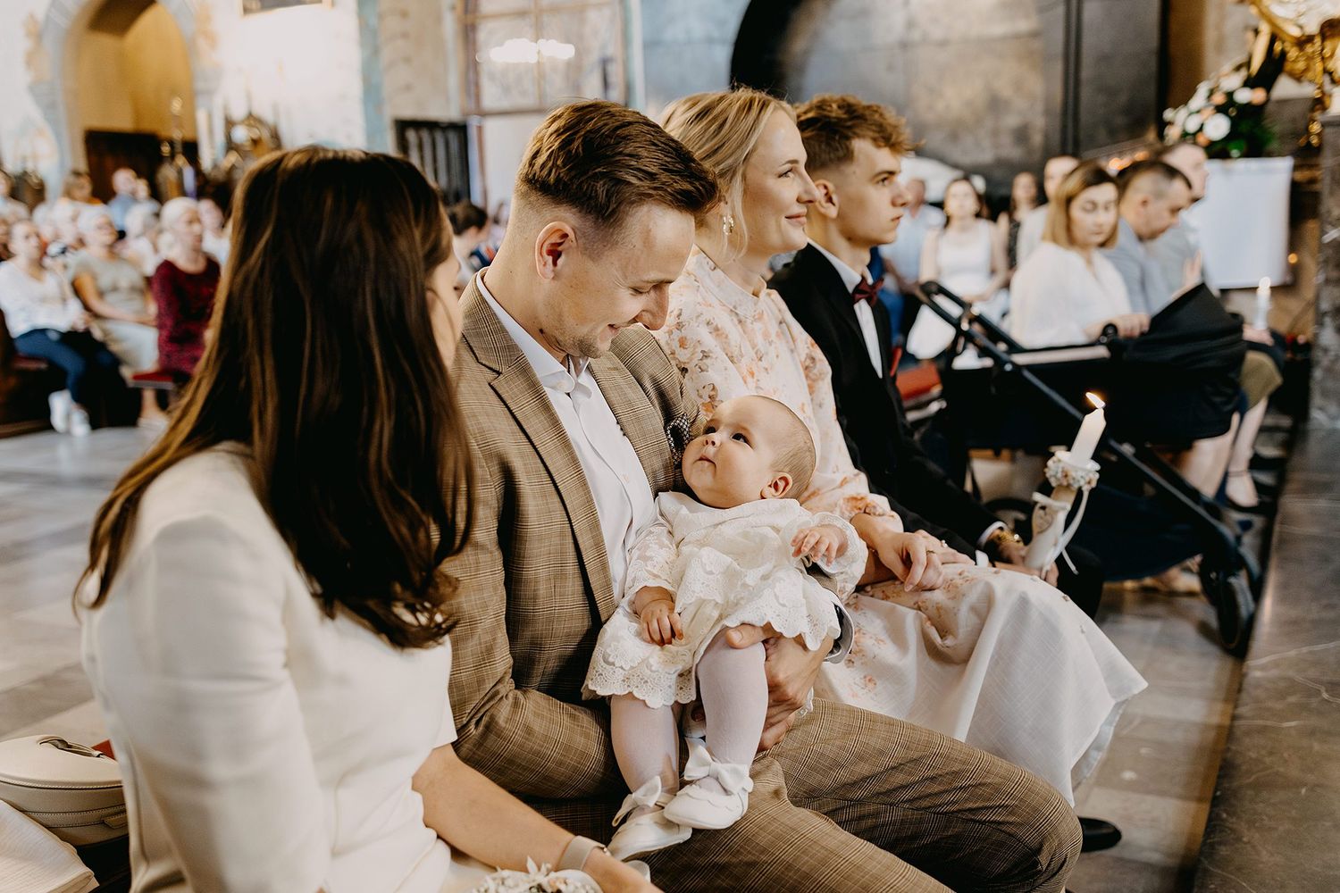 Family seated in church pew with baby in white christening dress during Catholic baptism ceremony.