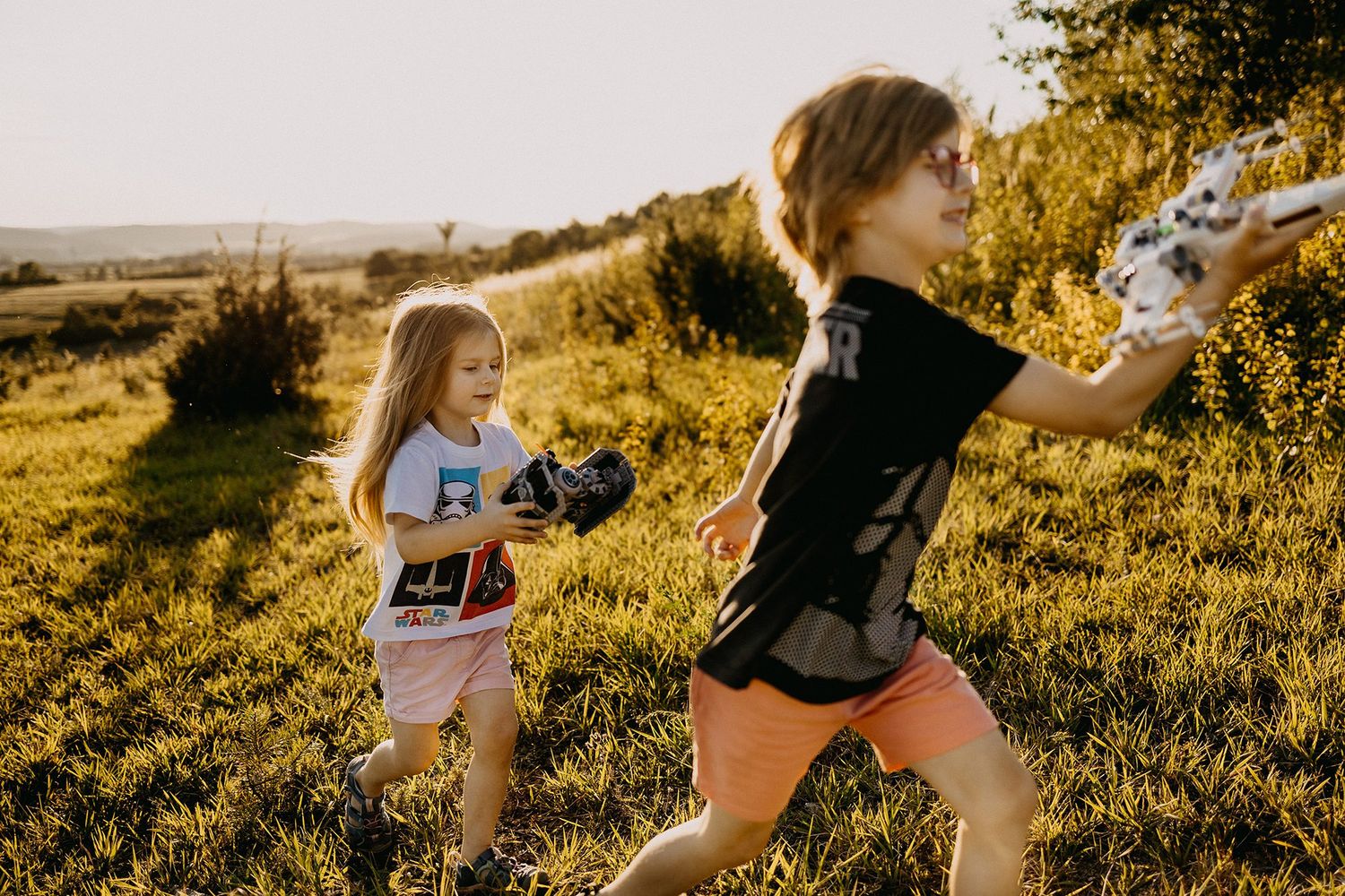 Two young girls playing with toy guns in a sunlit green field during golden hour outdoors.