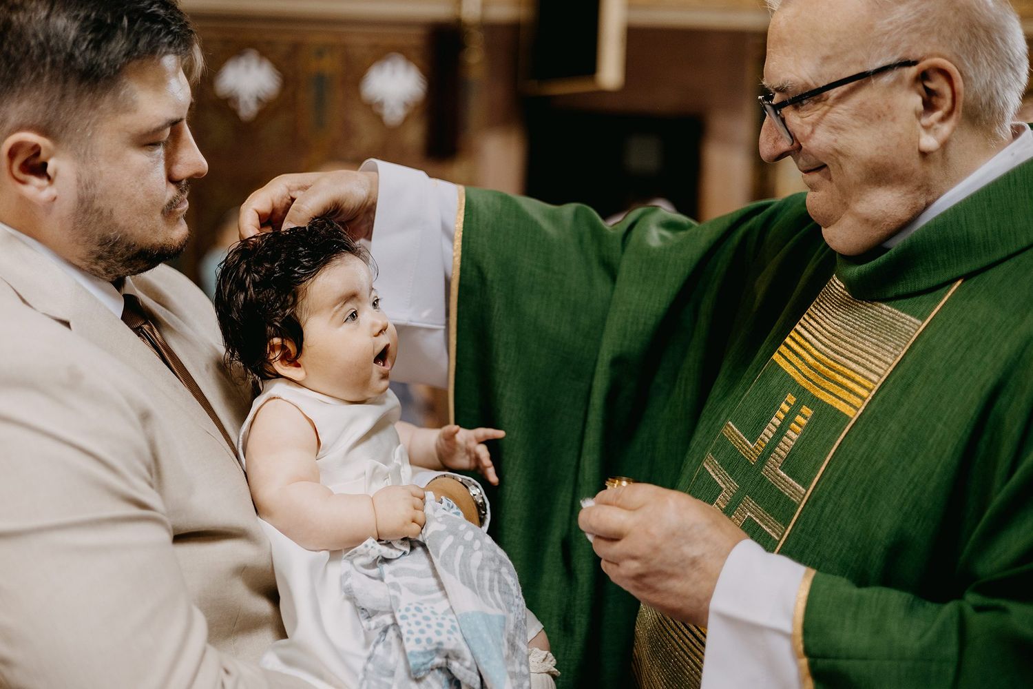 Catholic priest in green vestments performing baptism blessing on baby held by father in church.