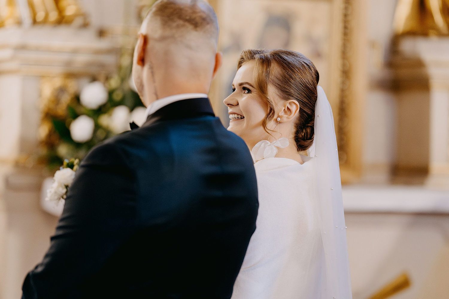 Bride in white veil smiling lovingly at groom during elegant church wedding ceremony.