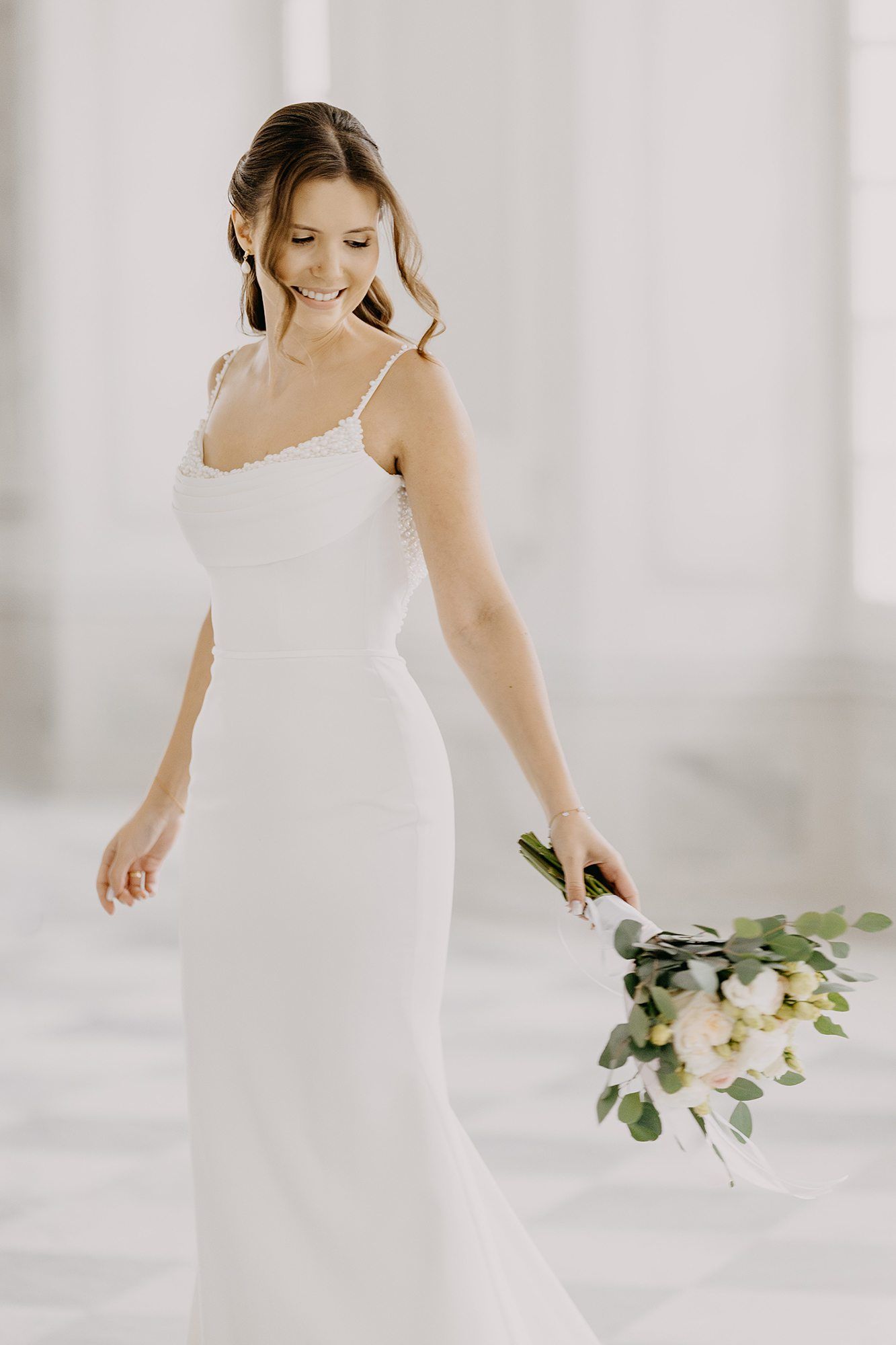 Smiling bride in sleek white spaghetti strap gown holding floral bouquet in bright setting.
