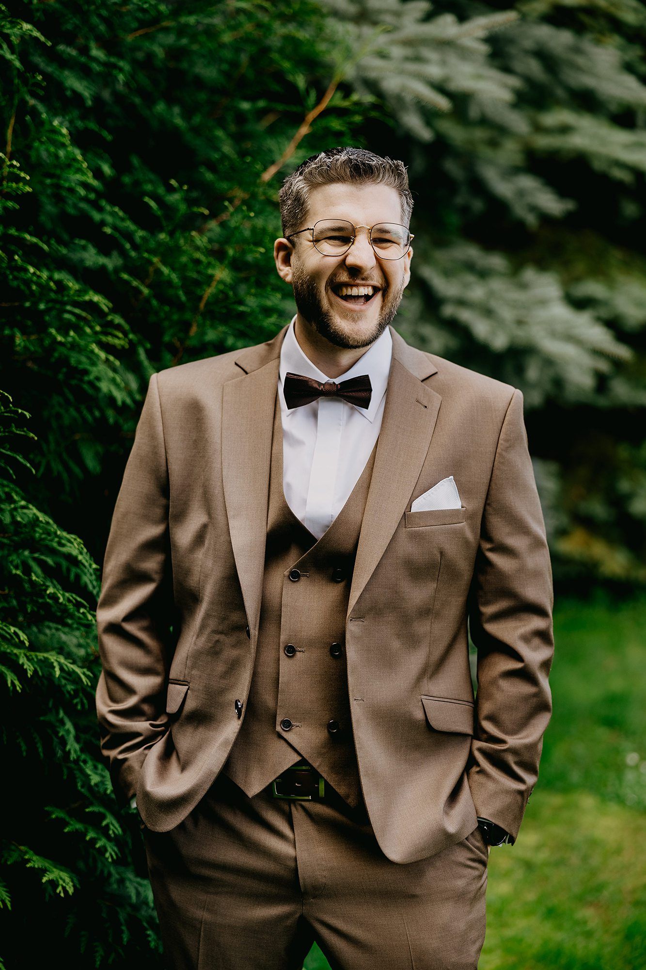 Smiling groom in tan three-piece suit with bow tie posing outdoors.