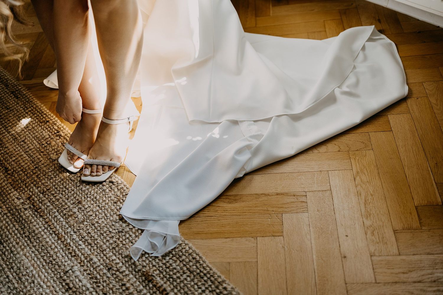 Bride adjusting white strappy heels on herringbone wood floor with flowing wedding dress train spread elegantly.