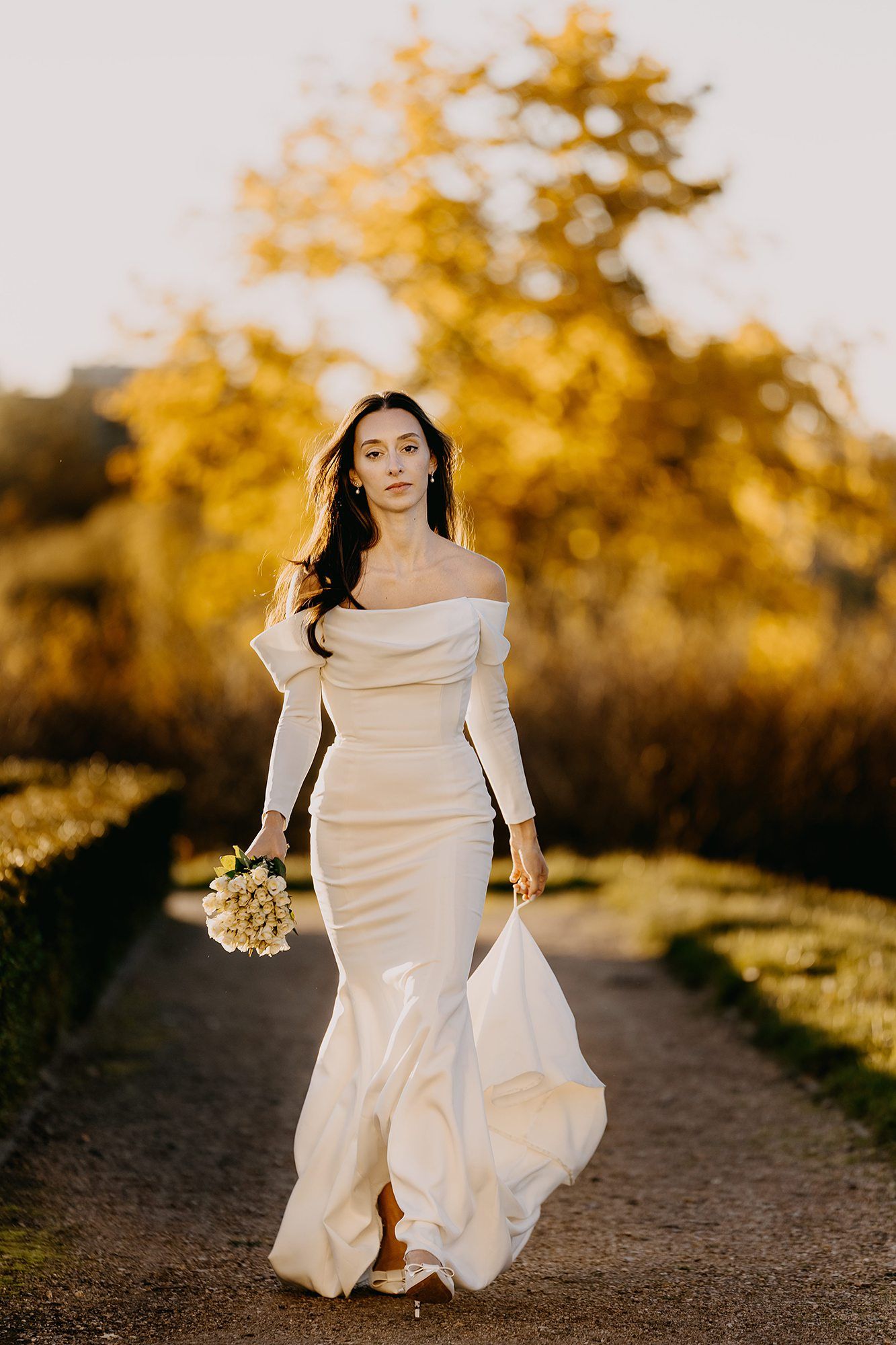 Elegant bride in off-shoulder white mermaid gown walks garden path amid golden autumn foliage.