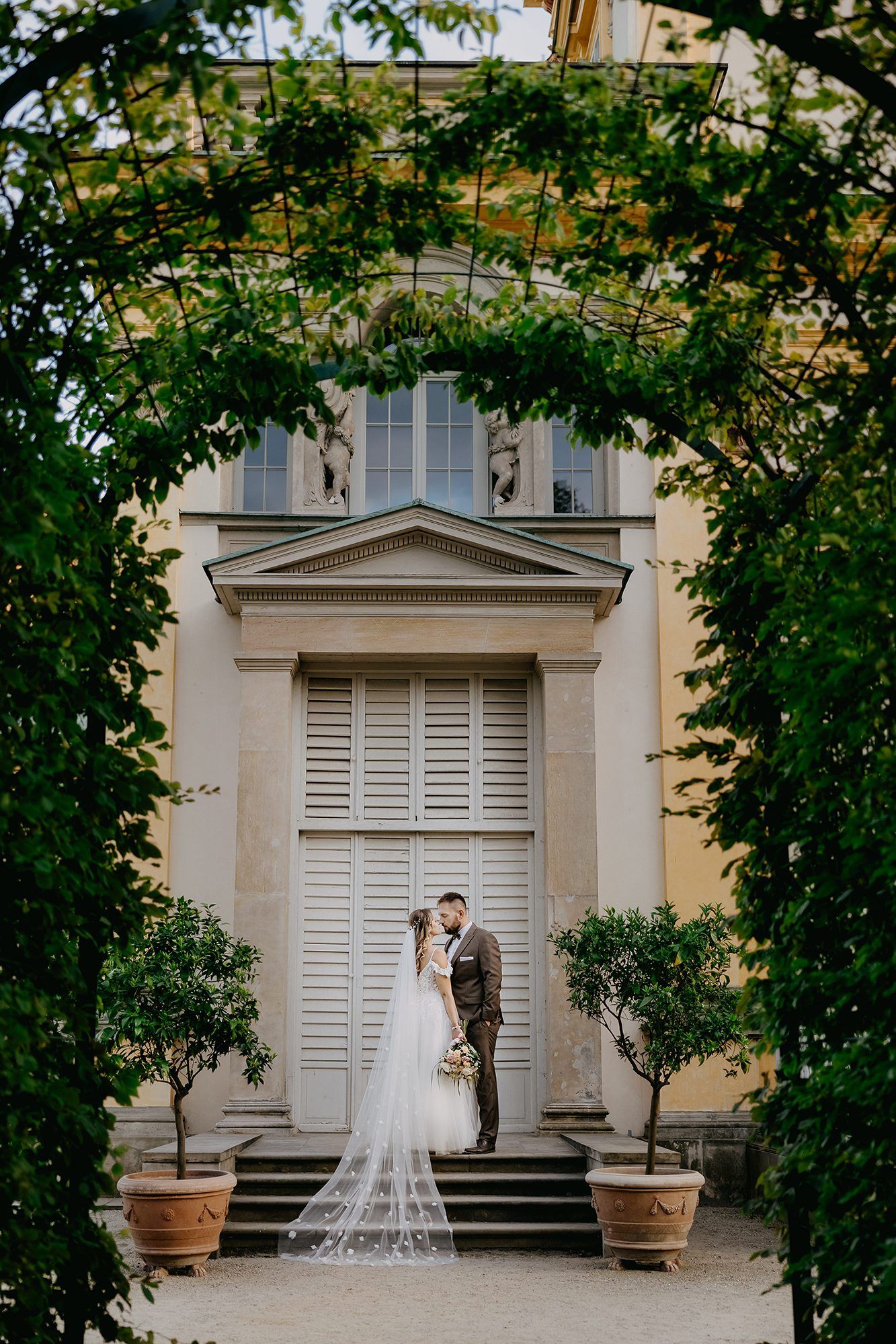 Bride and groom kissing framed by green archway before grand neoclassical building with long veil.