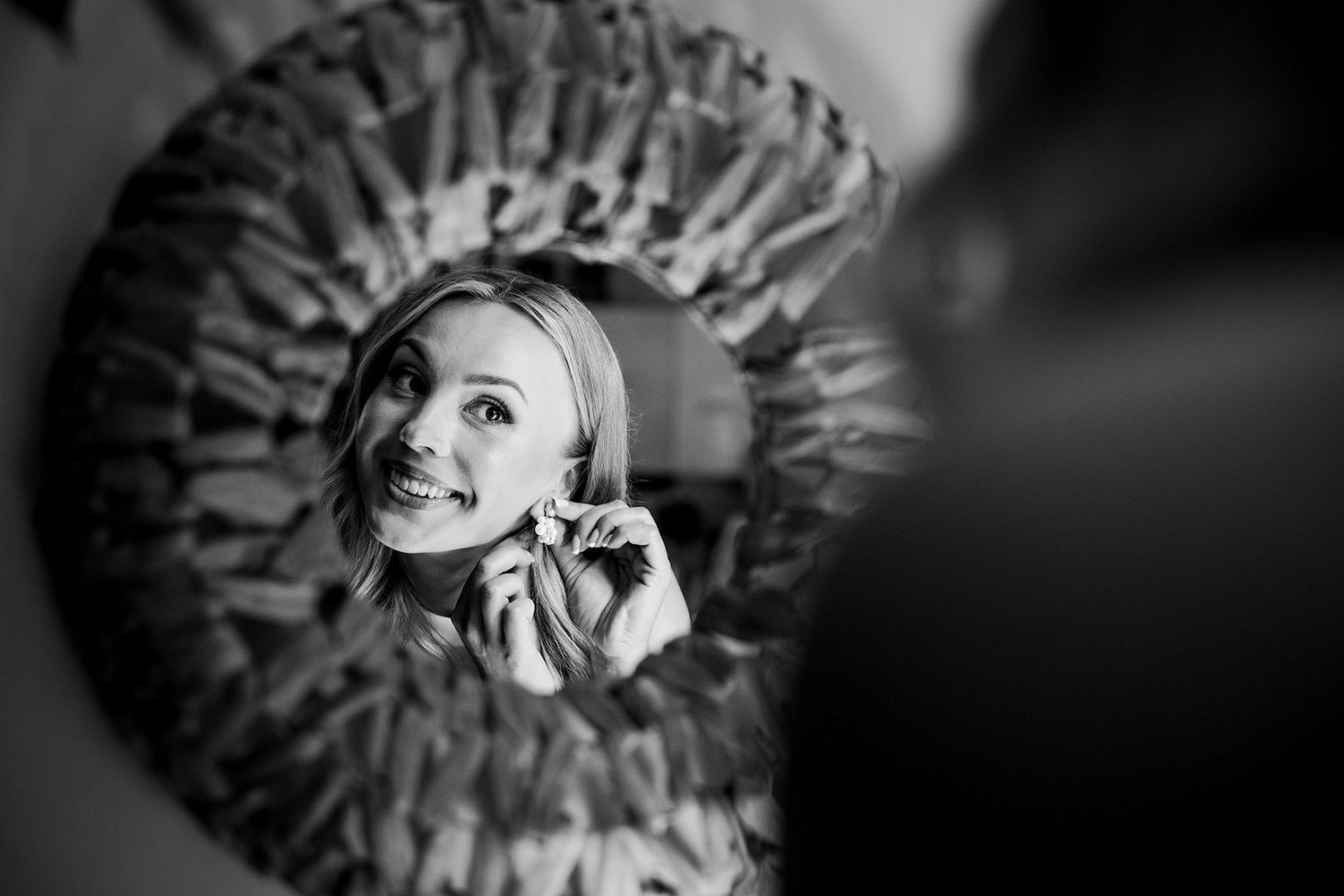 Black and white reflection of a smiling bride putting on earrings in a decorative round mirror.