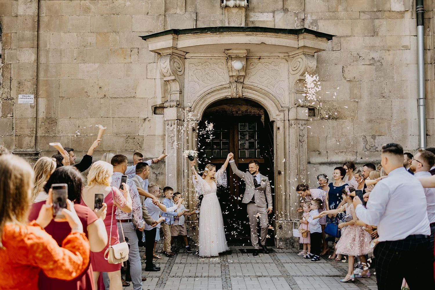 A joyful bride and groom exit a historic stone church as guests shower them with confetti in celebration.
