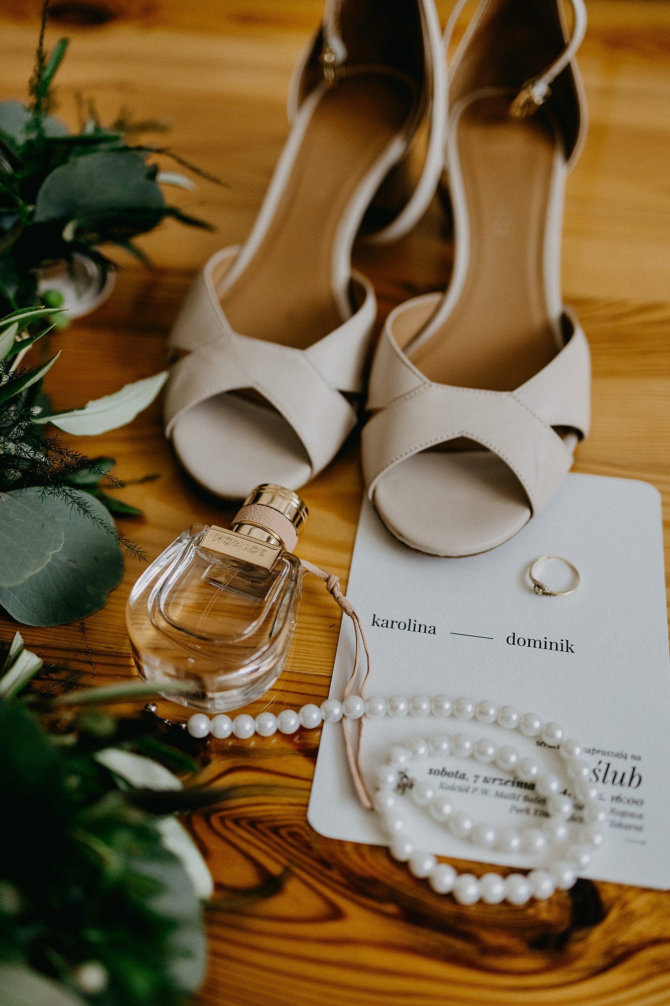 Wedding flat lay featuring nude heels, pearl bracelet, perfume, ring, and invitation card on wood.