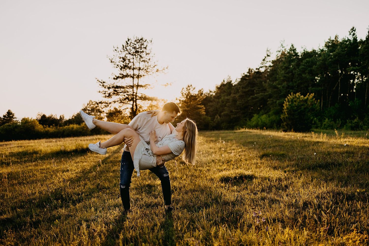 A man lifts and spins a woman in a golden sunset field surrounded by trees.