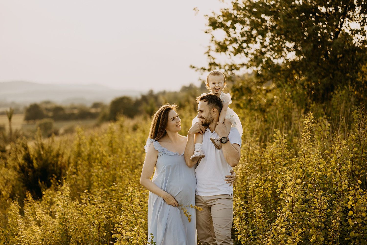 Happy family with toddler on shoulders standing in golden wildflower field at sunset.