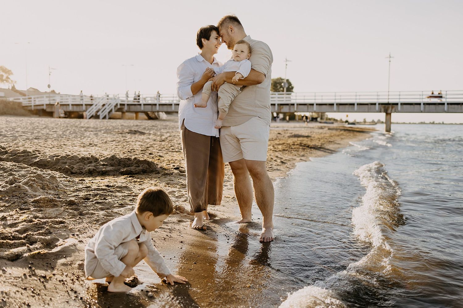 A happy family of four enjoys a golden sunset beach outing with a pier visible in the background.