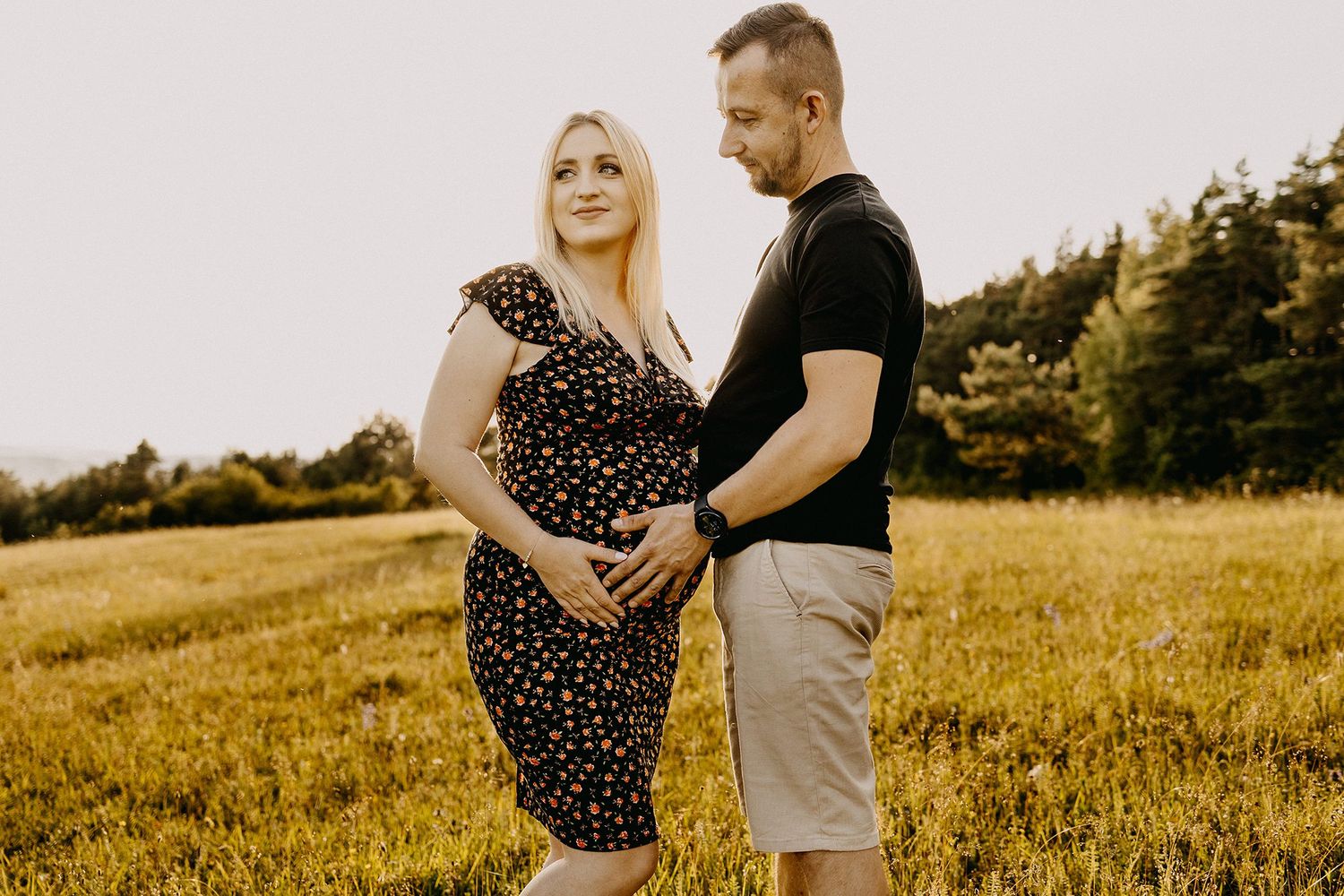 Expecting couple standing in sunny meadow, man tenderly touching pregnant womans baby bump, golden hour light.