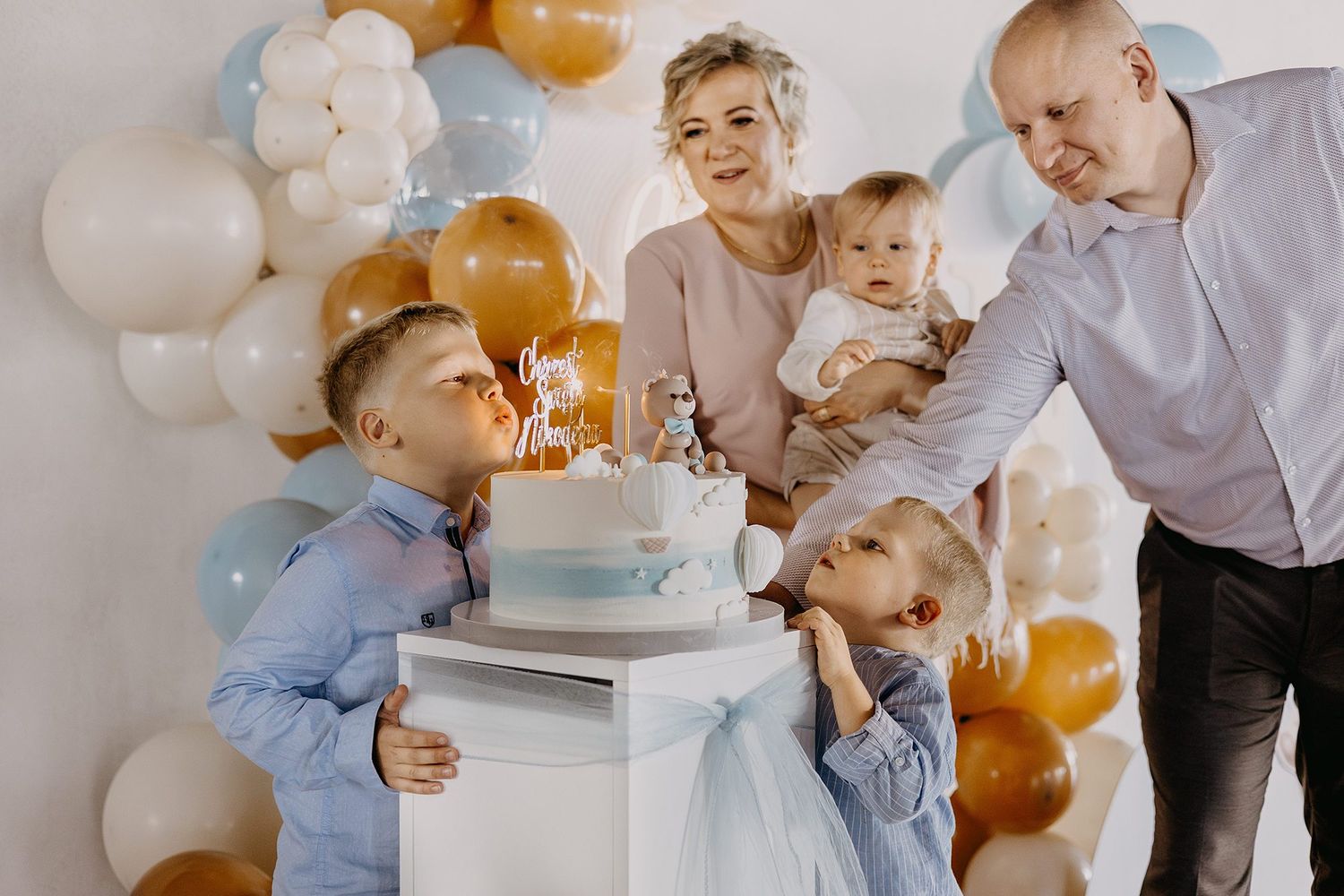 Family celebrating baby first birthday with blue cake and balloon decorations.