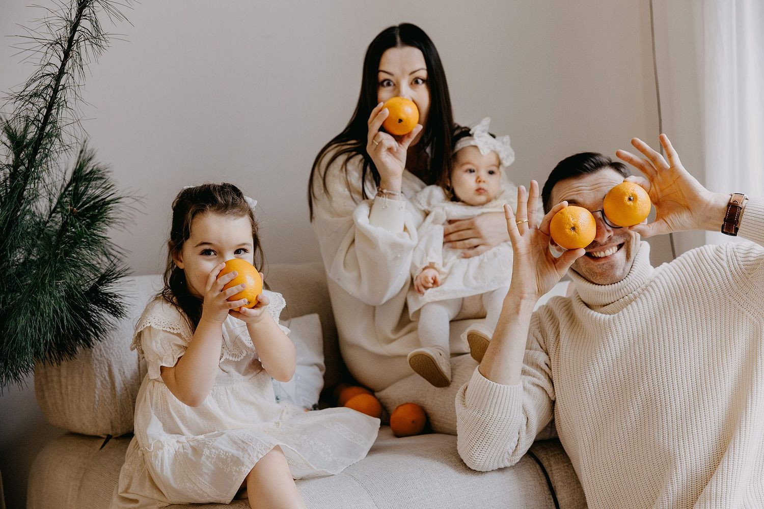 A cheerful family of four playfully holds oranges to their faces while sitting together in festive white outfits.