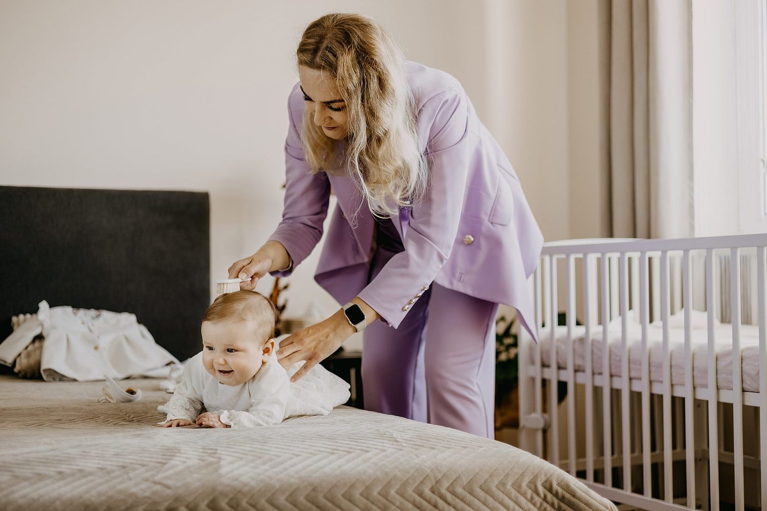 A mother in a lavender suit tends to her baby on a bed next to a white crib.