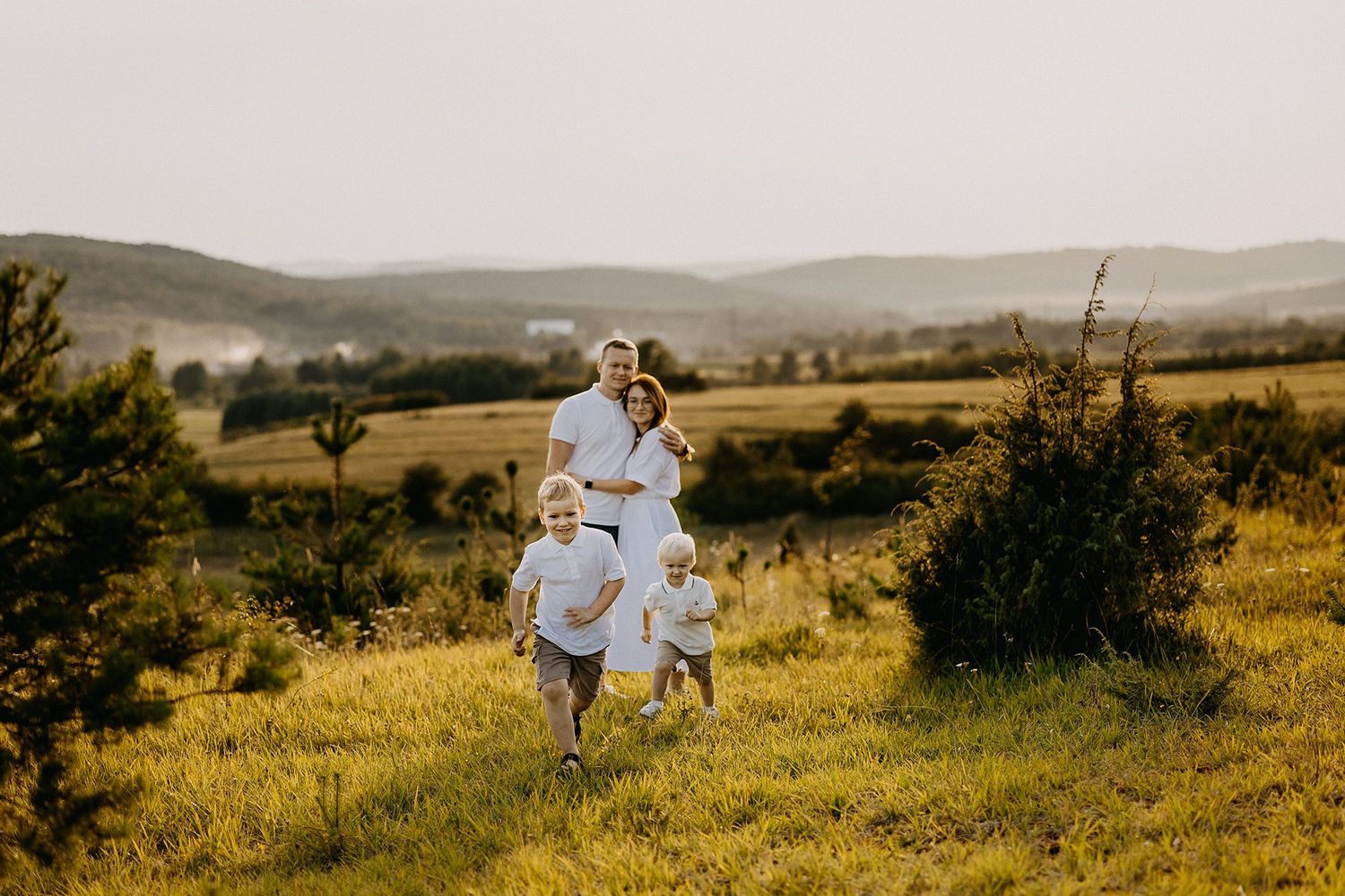 Family of four in white outfits walking together through a scenic green countryside at golden hour.