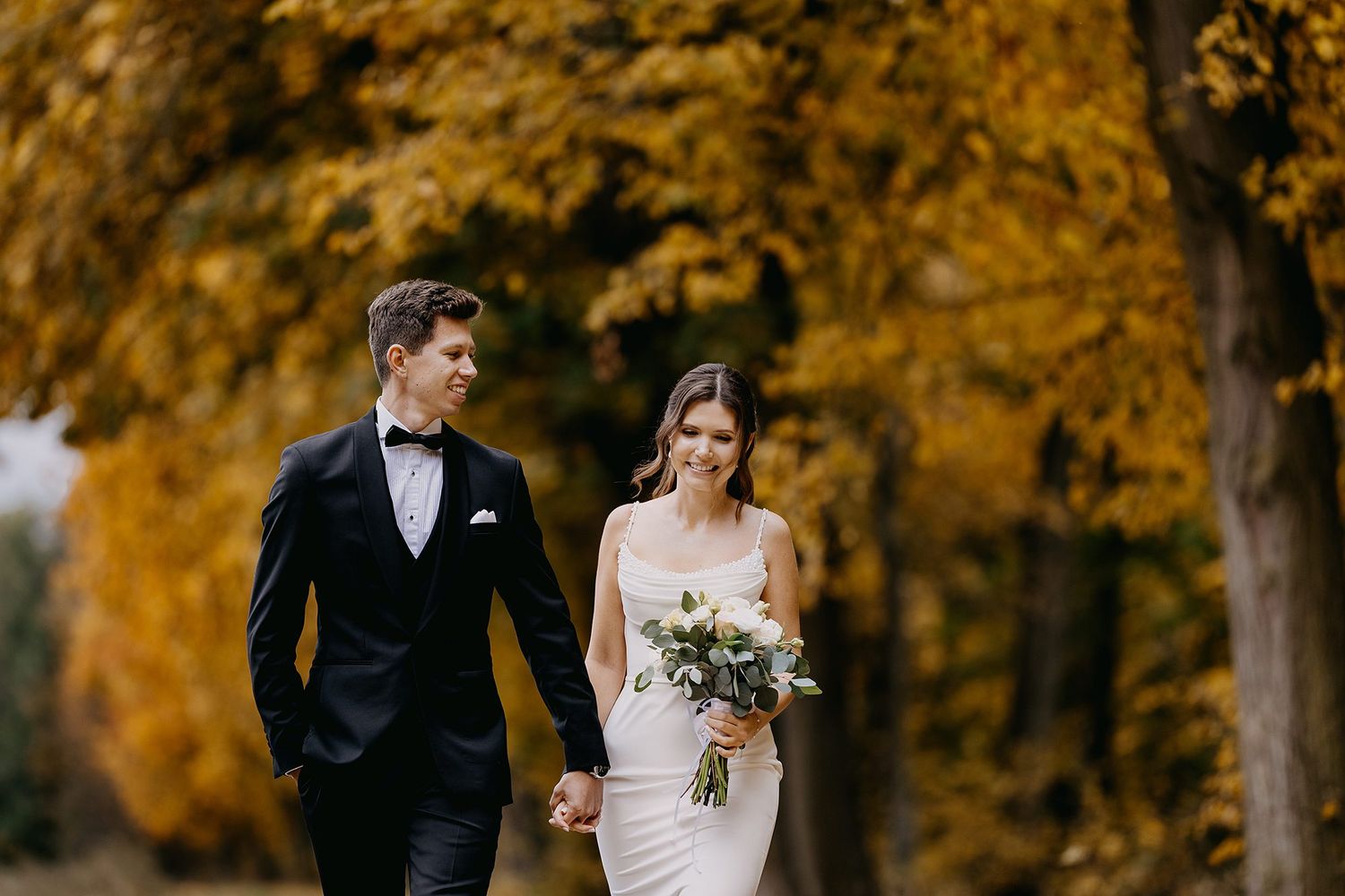 Bride and groom walking hand in hand through a path lined with golden autumn trees.