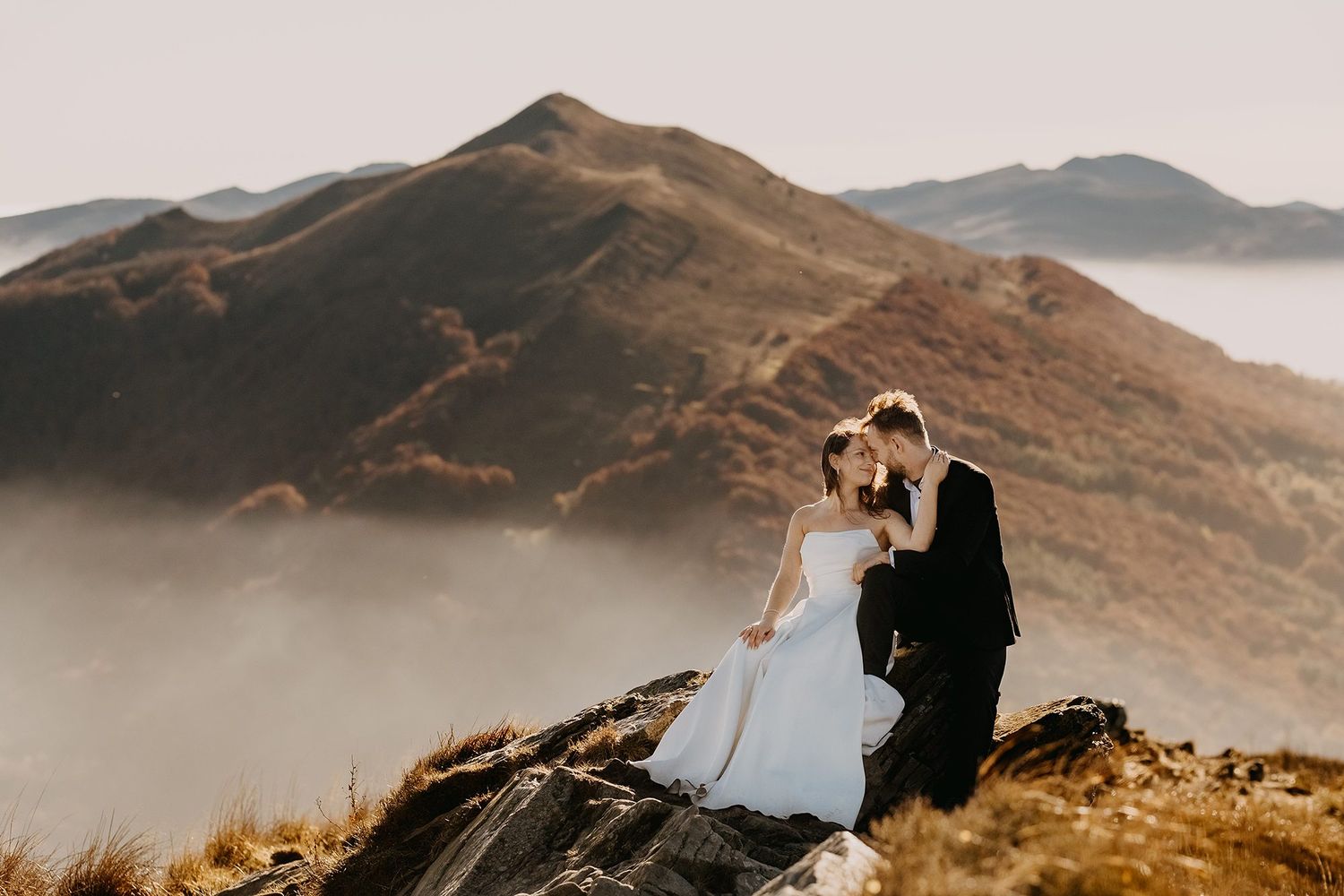 Romantic bride and groom sitting on a rocky mountain peak with misty hills and clouds below.
