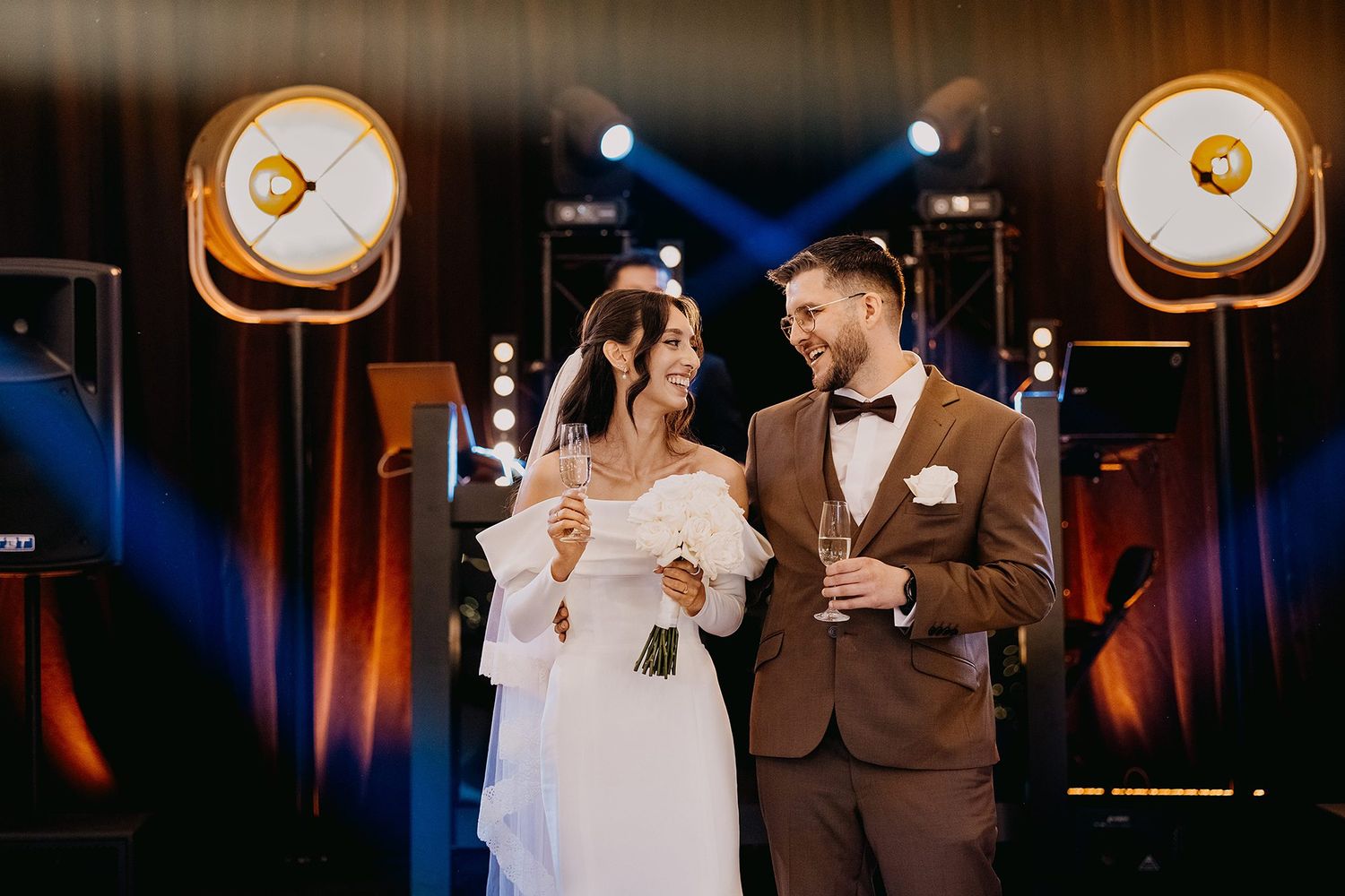 Bride and groom share champagne toast under dramatic stage lights at elegant wedding reception venue.