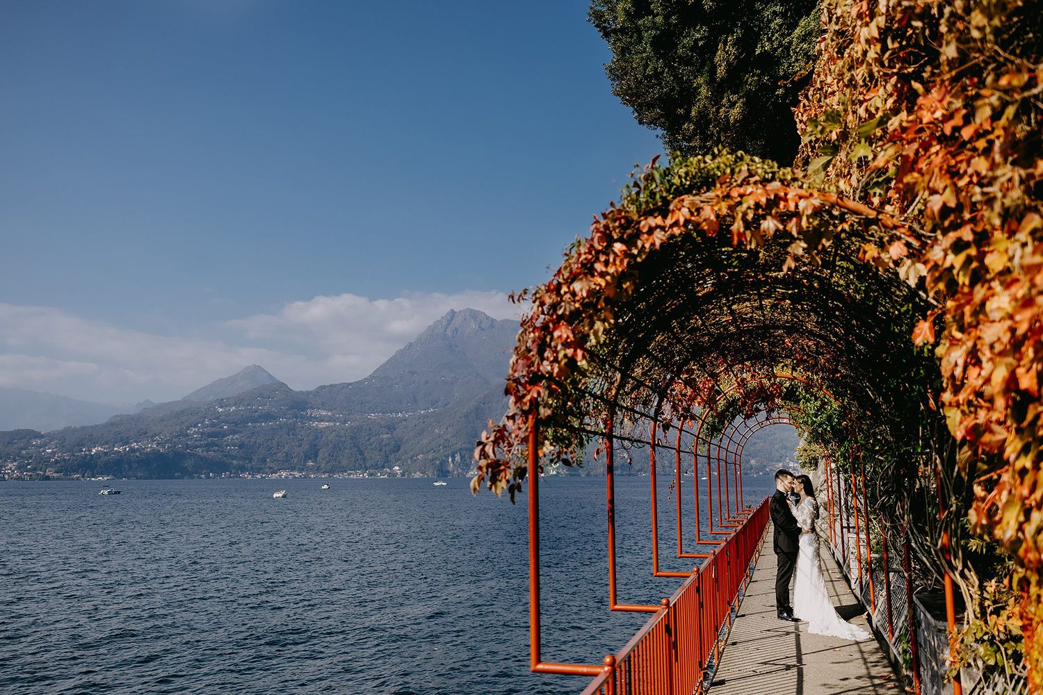 Wedding couple embraces under autumn ivy arches on a lakeside promenade with mountain views.