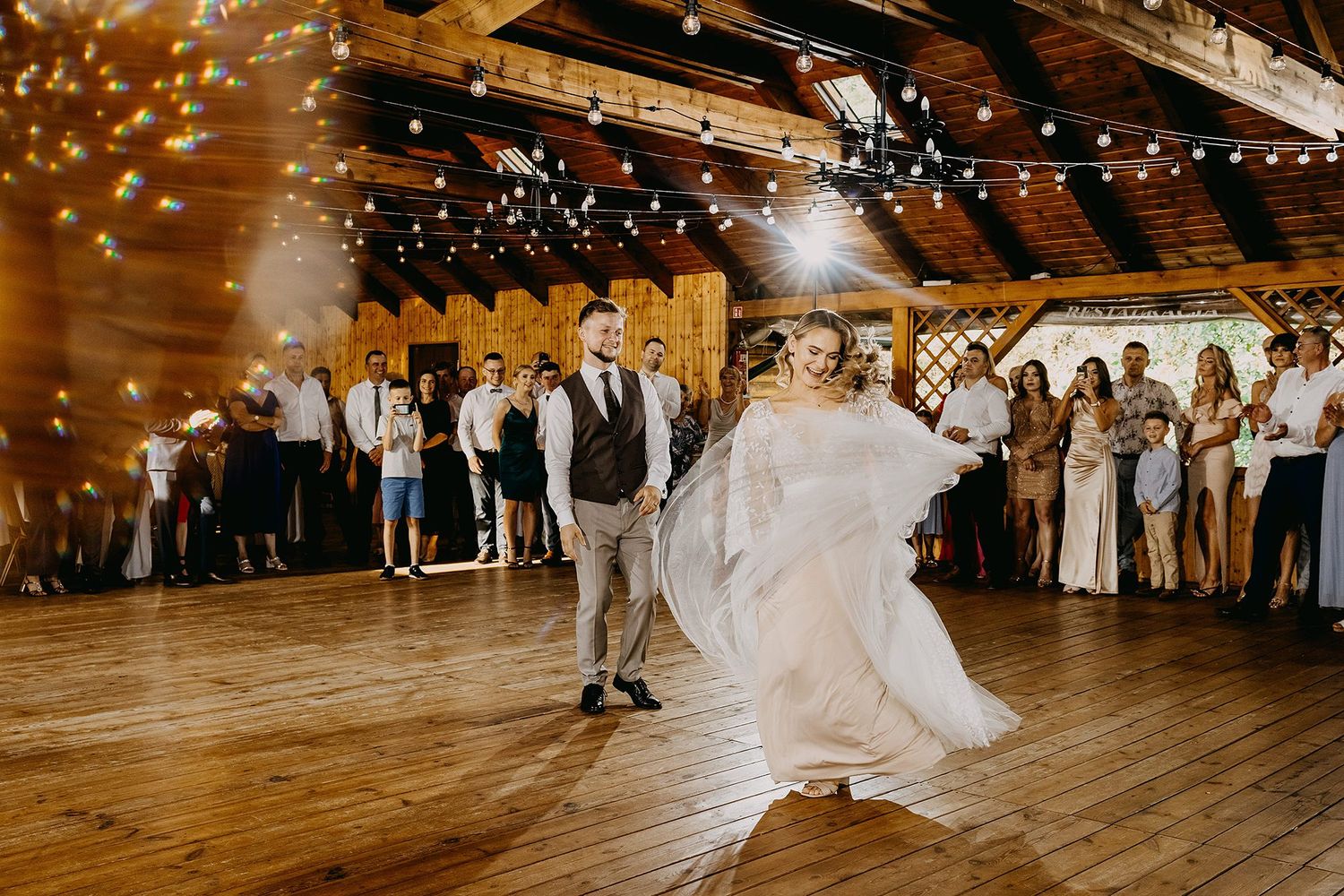 Bride and groom share first dance on wooden floor in rustic barn venue with string lights.