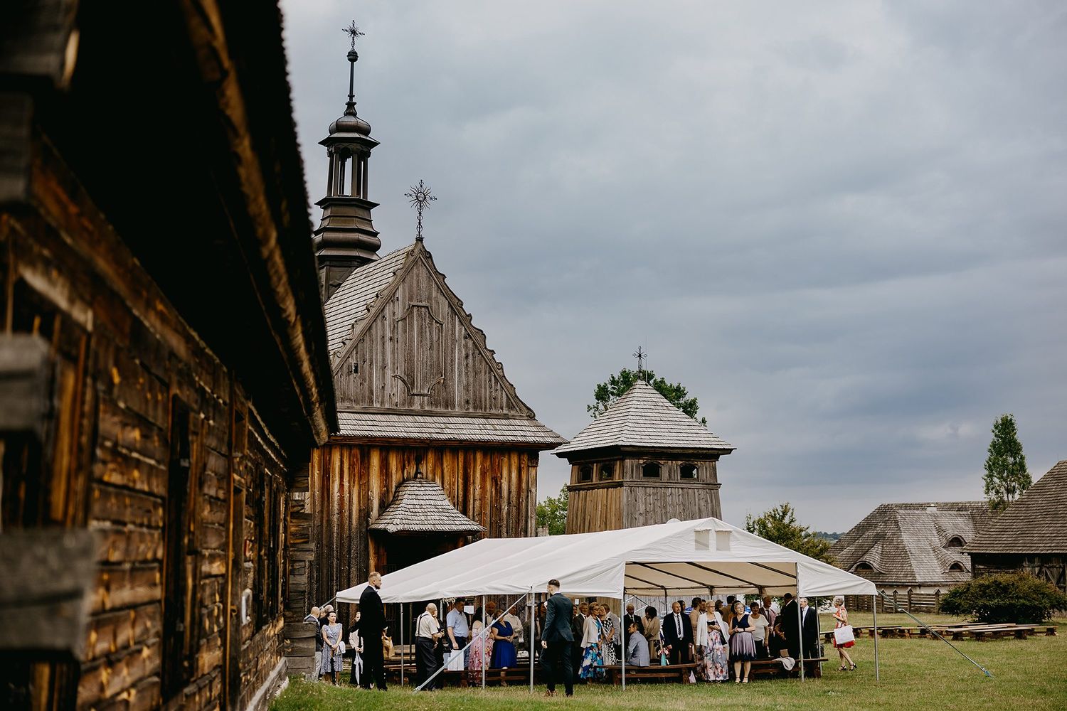 Outdoor wedding reception with white marquee tent near historic wooden church and bell tower on cloudy day.