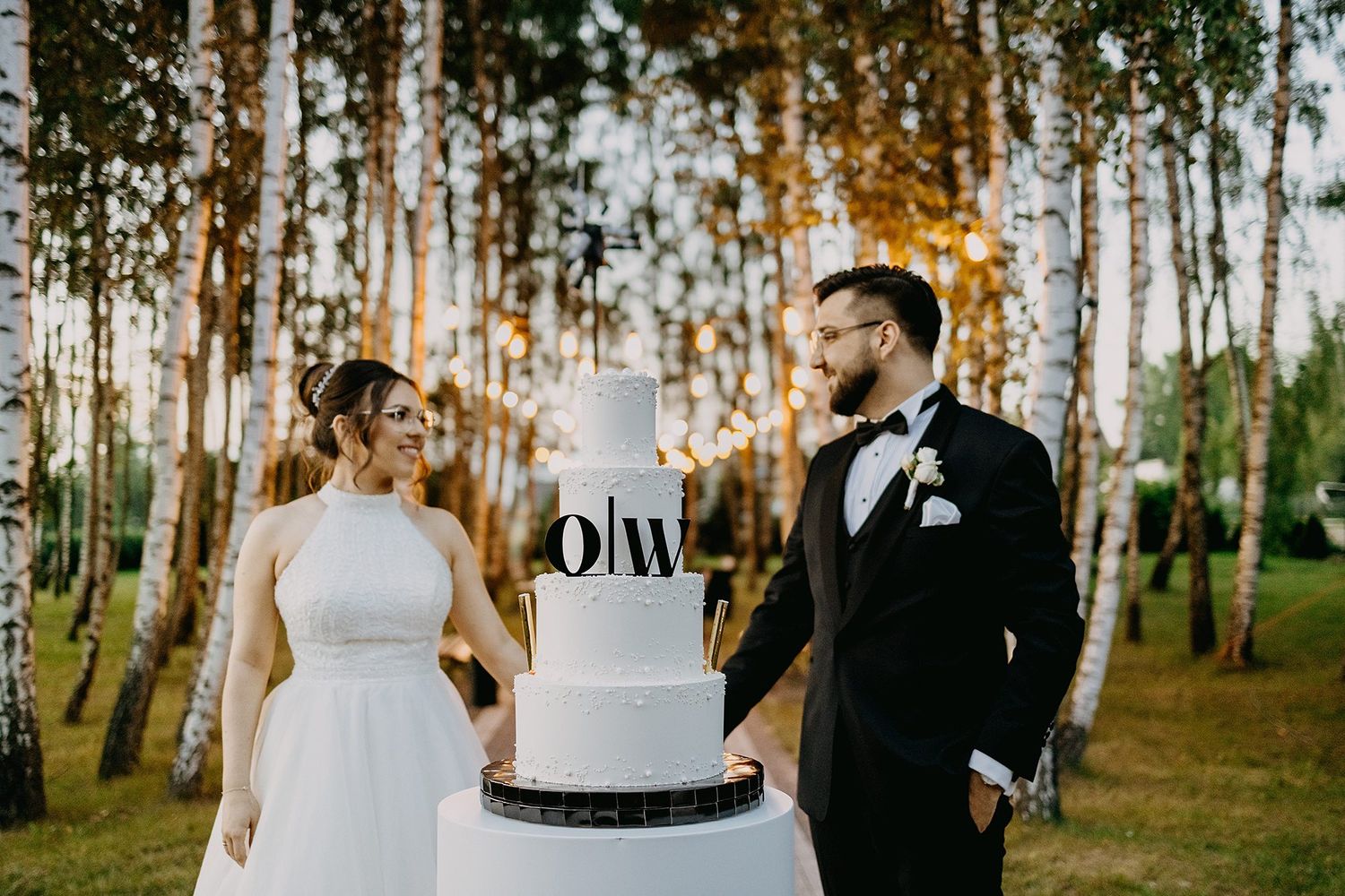 Bride and groom smiling beside a white four-tier wedding cake with monogram topper in a birch tree forest.